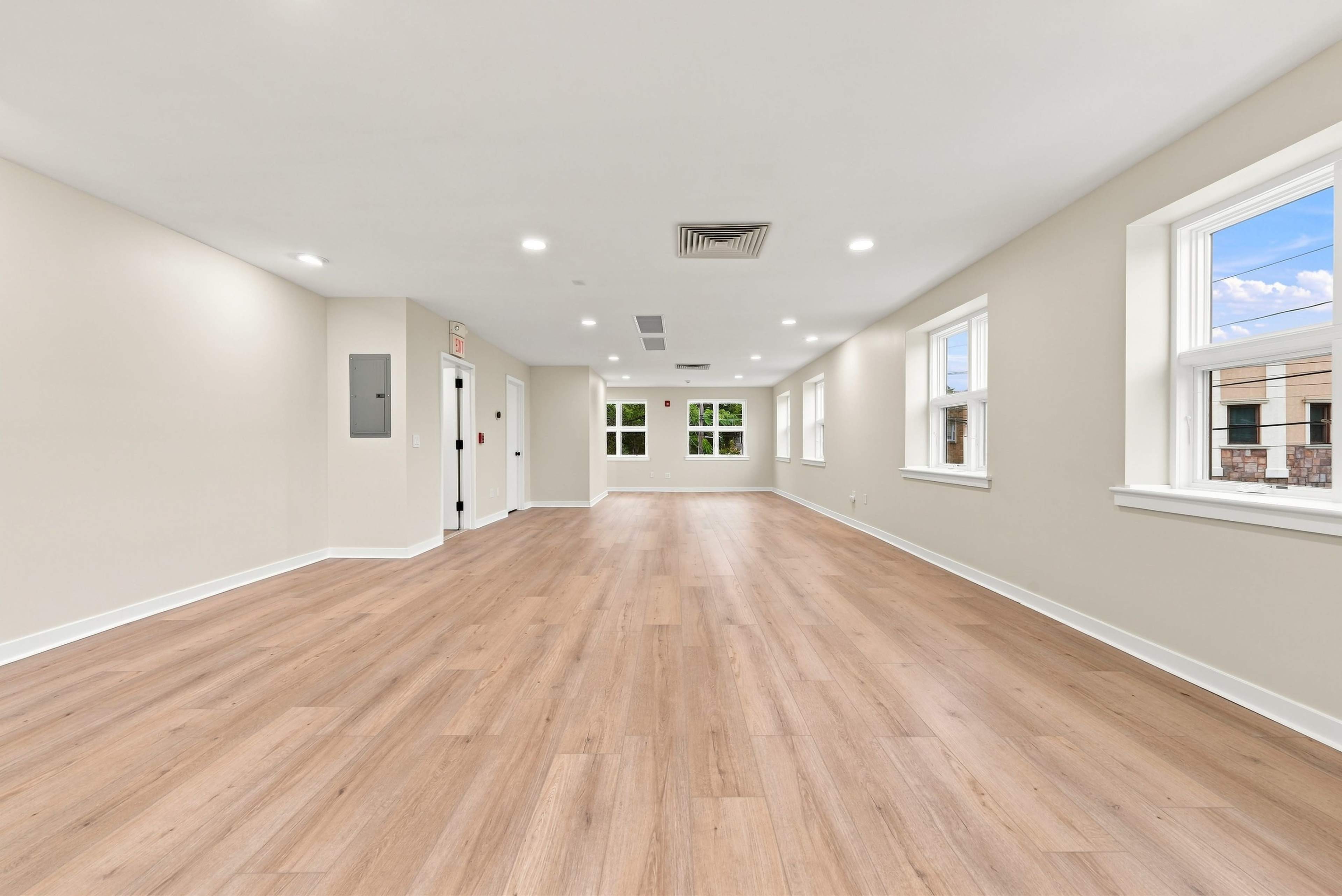 The image shows a spacious, empty room with light wood flooring, plain walls, and several windows providing natural light.