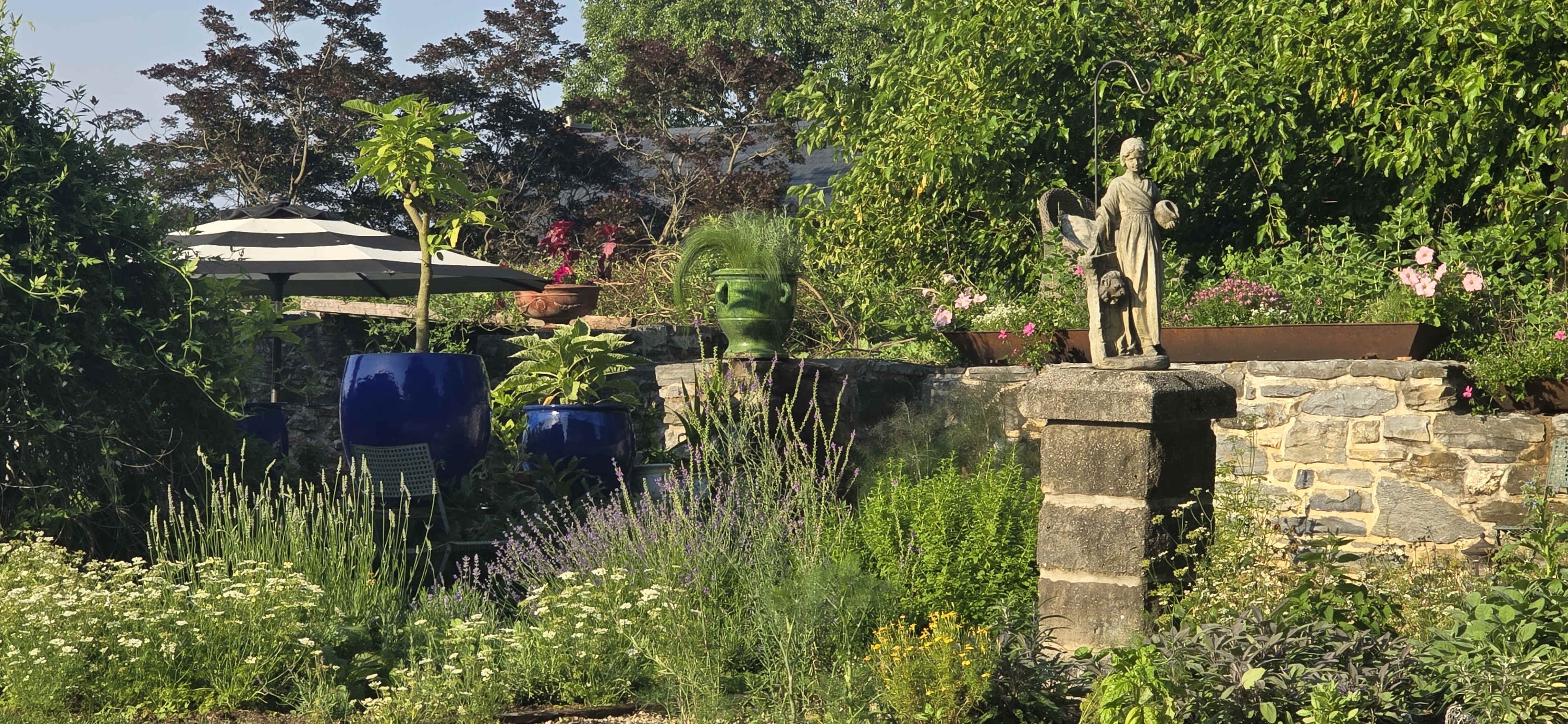 A well-kept garden features a statue on a stone pedestal surrounded by various plants, flowers, and a shaded seating area under an umbrella.