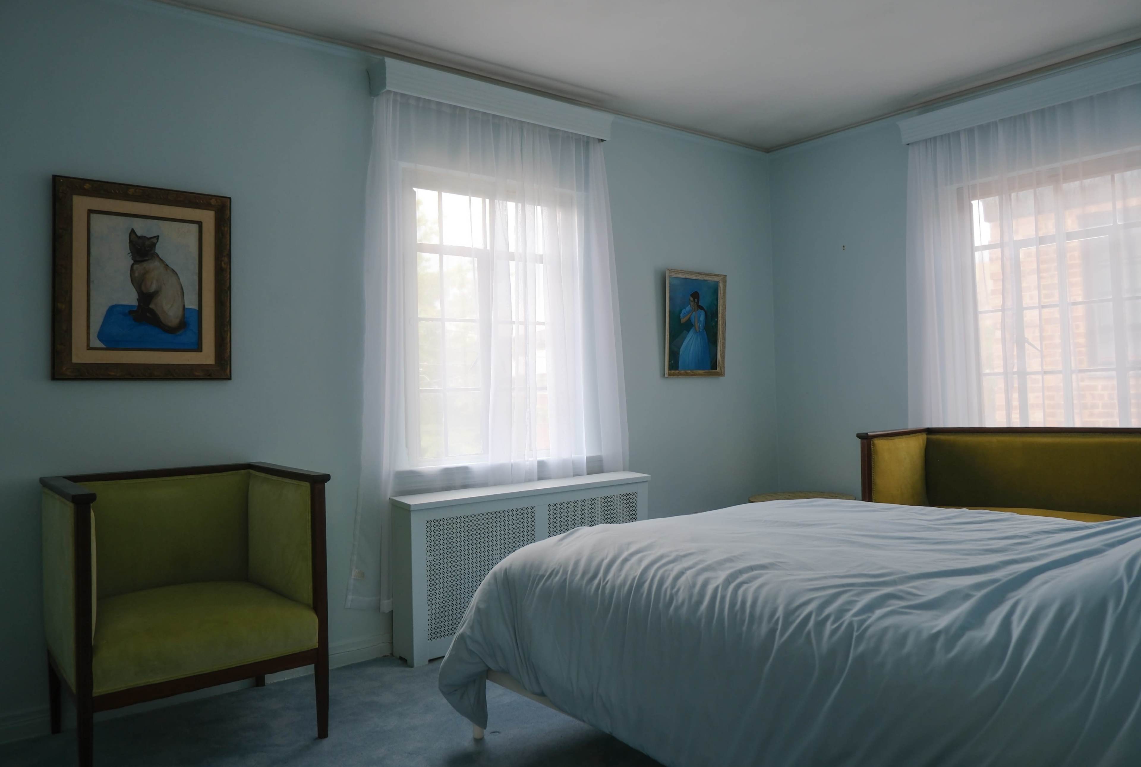 The image shows a light-colored bedroom furnished with a bed, two chairs, and framed artwork on the walls.