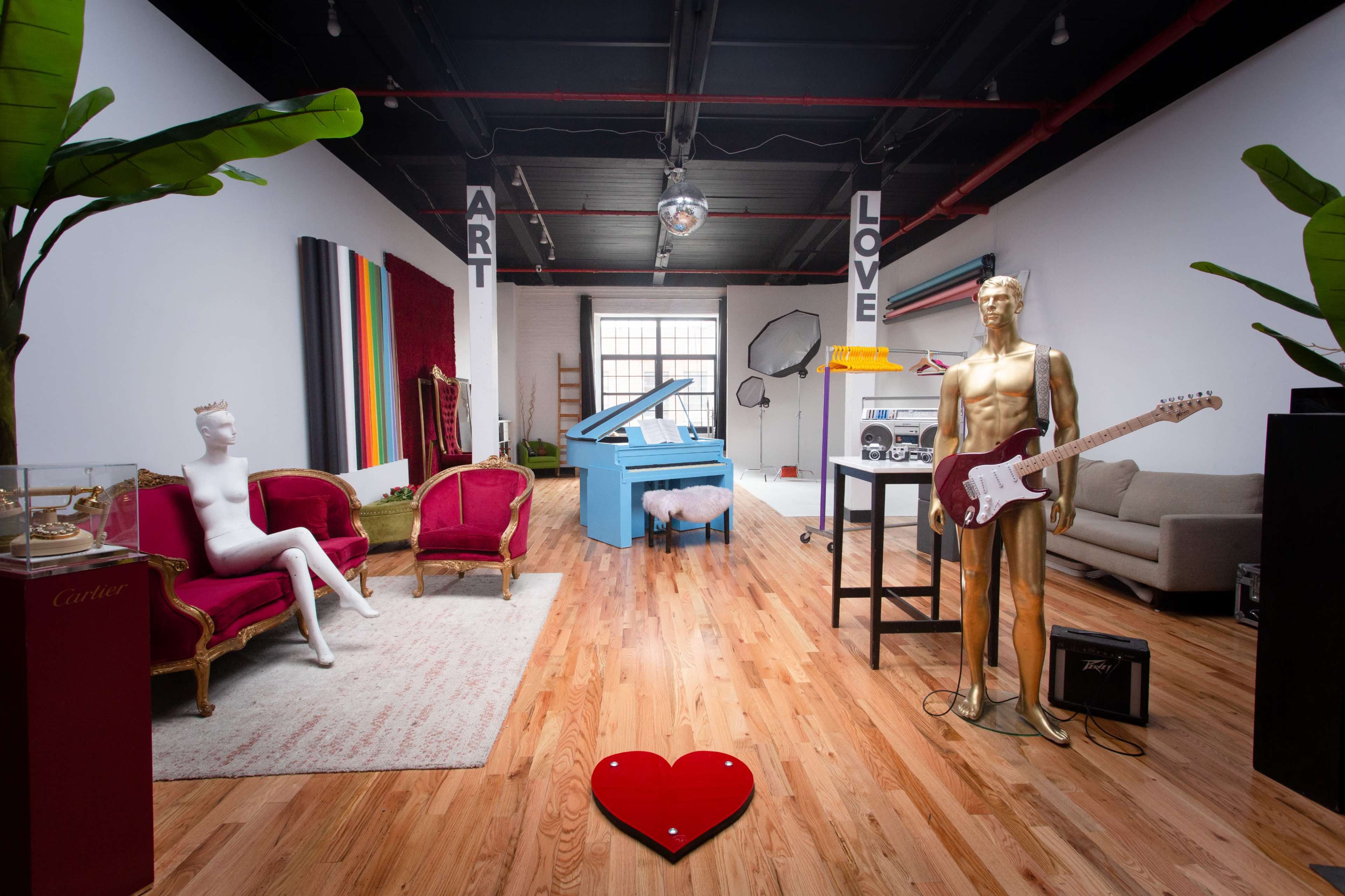 The image shows a spacious art studio featuring a blue piano, gold mannequin with a guitar, plush seating, and decorative elements like a heart-shaped design on the floor.