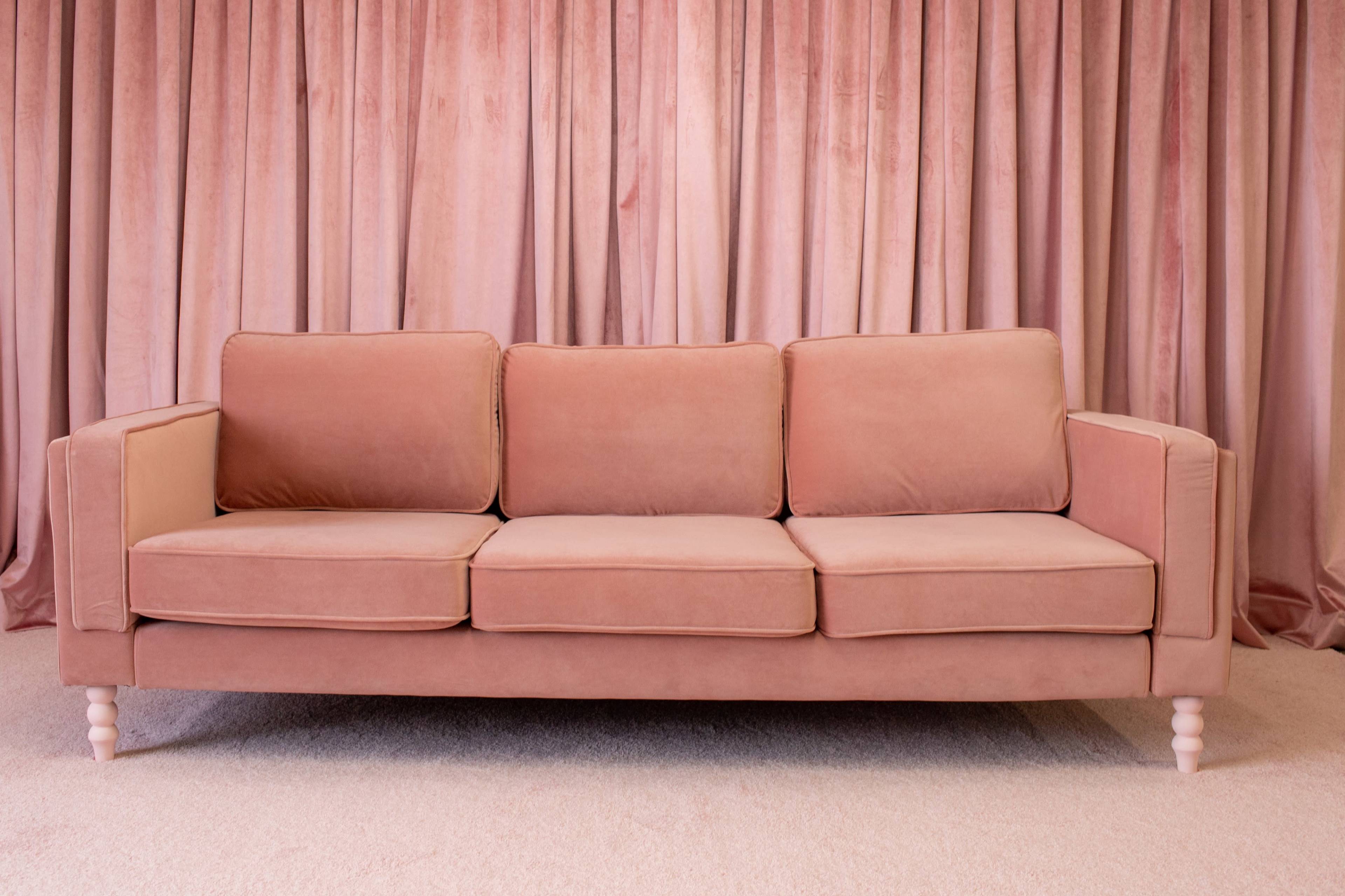 A pink velvet sofa is positioned in front of a matching curtain backdrop in a well-lit room.