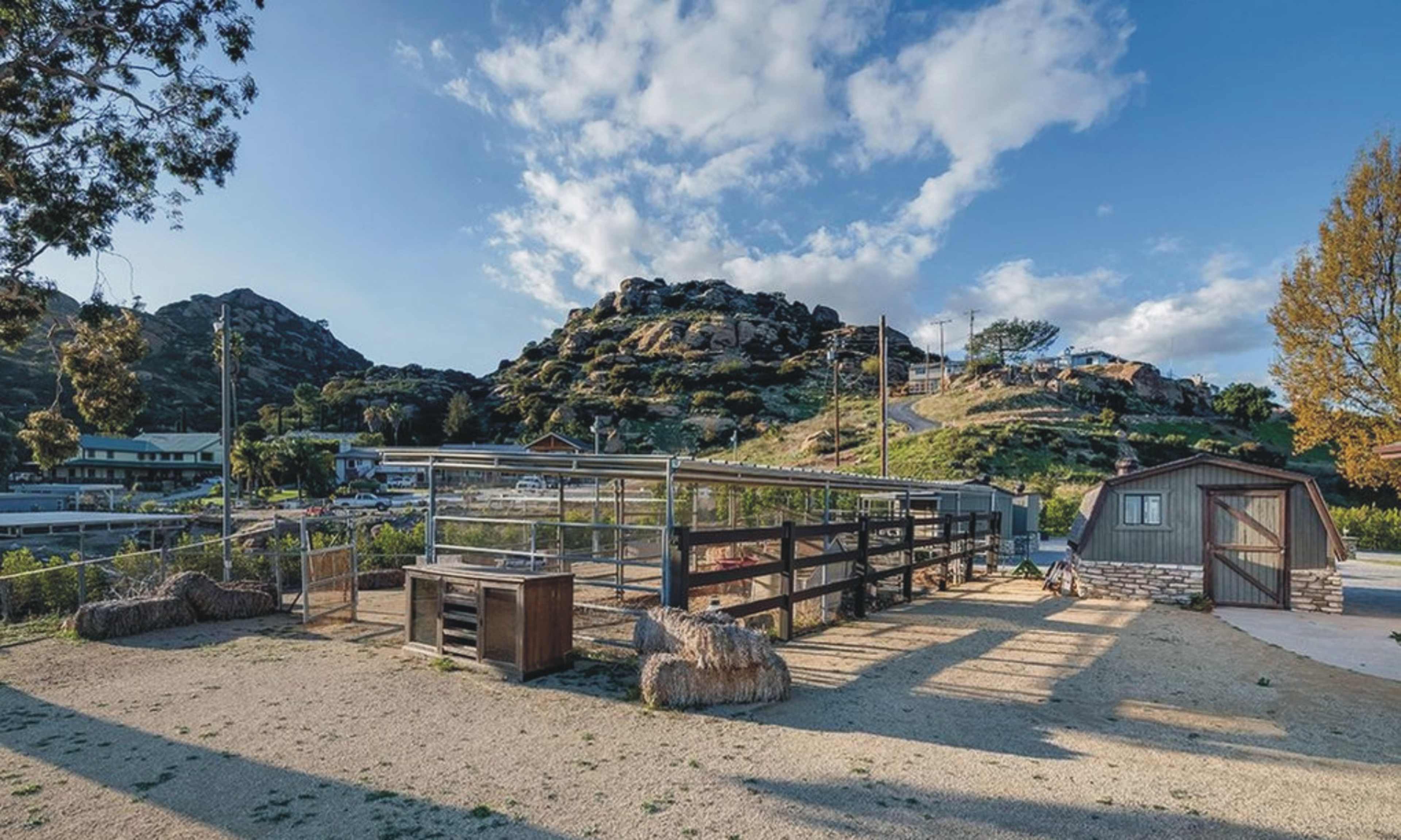 The scene features a fenced pasture area with animal enclosures, a small wooden shed, and rocky hills in the background under a partly cloudy sky.
