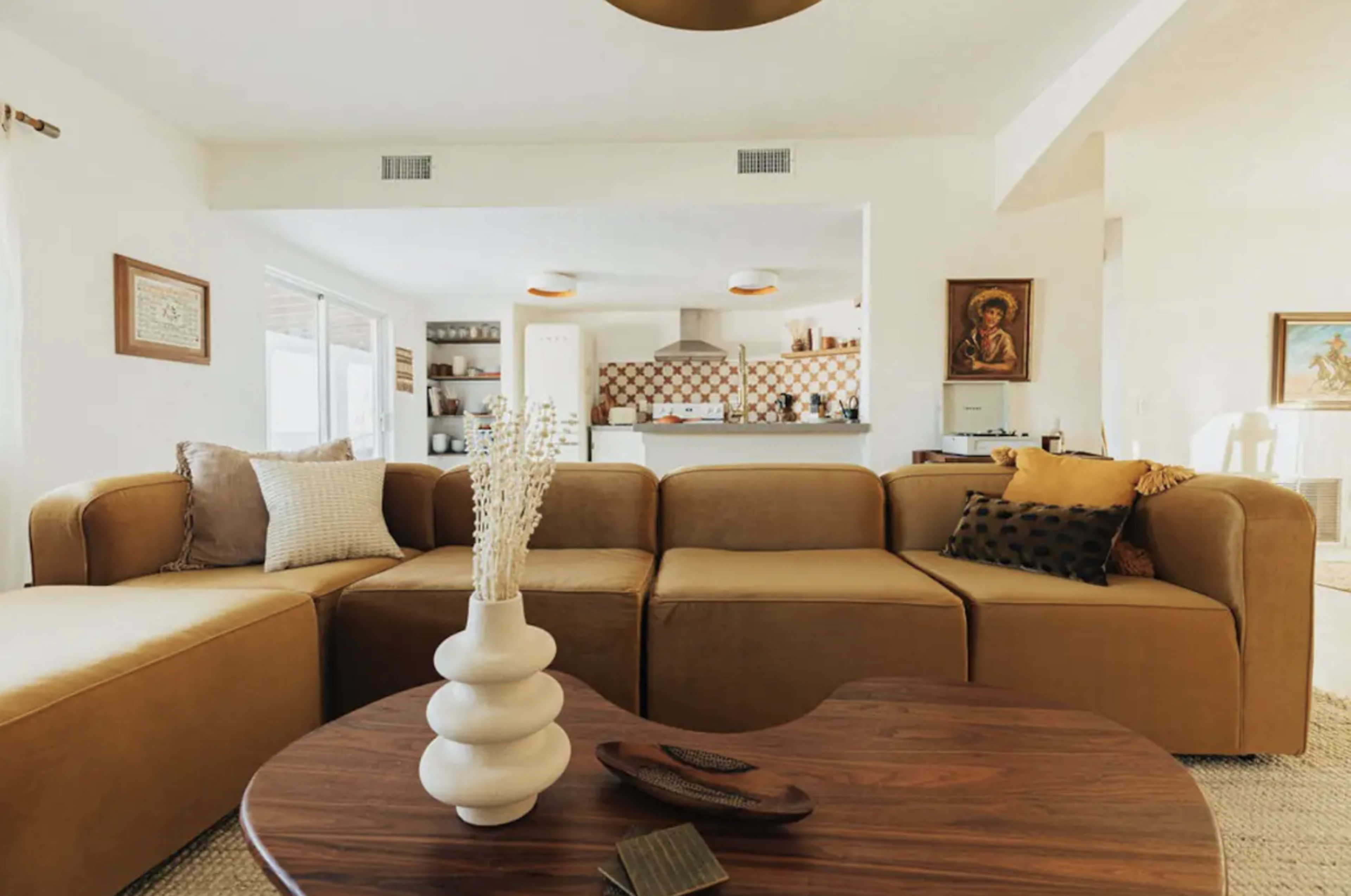 The living room features a large, L-shaped brown sectional sofa and a round wooden coffee table with decorative items on it, set against a backdrop of a kitchen area.