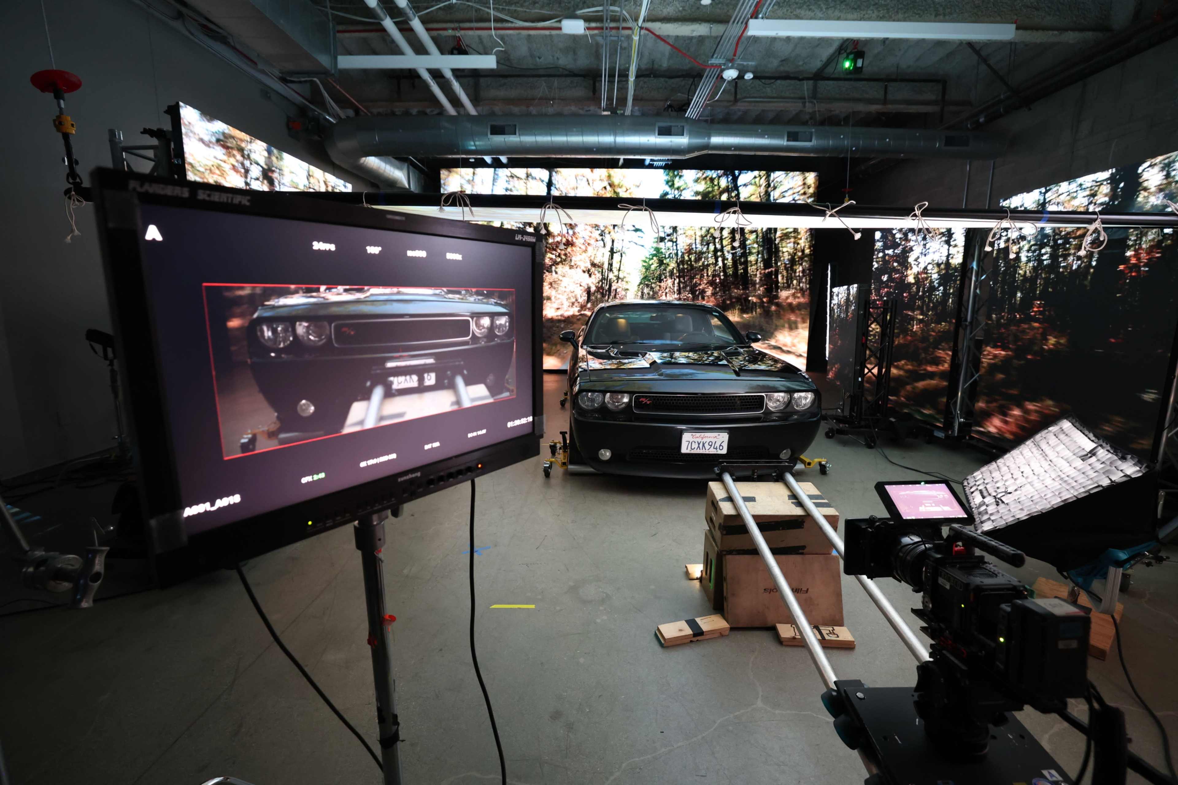 A black car is positioned on a set with a digital screen displaying a forest backdrop, accompanied by camera equipment and monitors for filming.