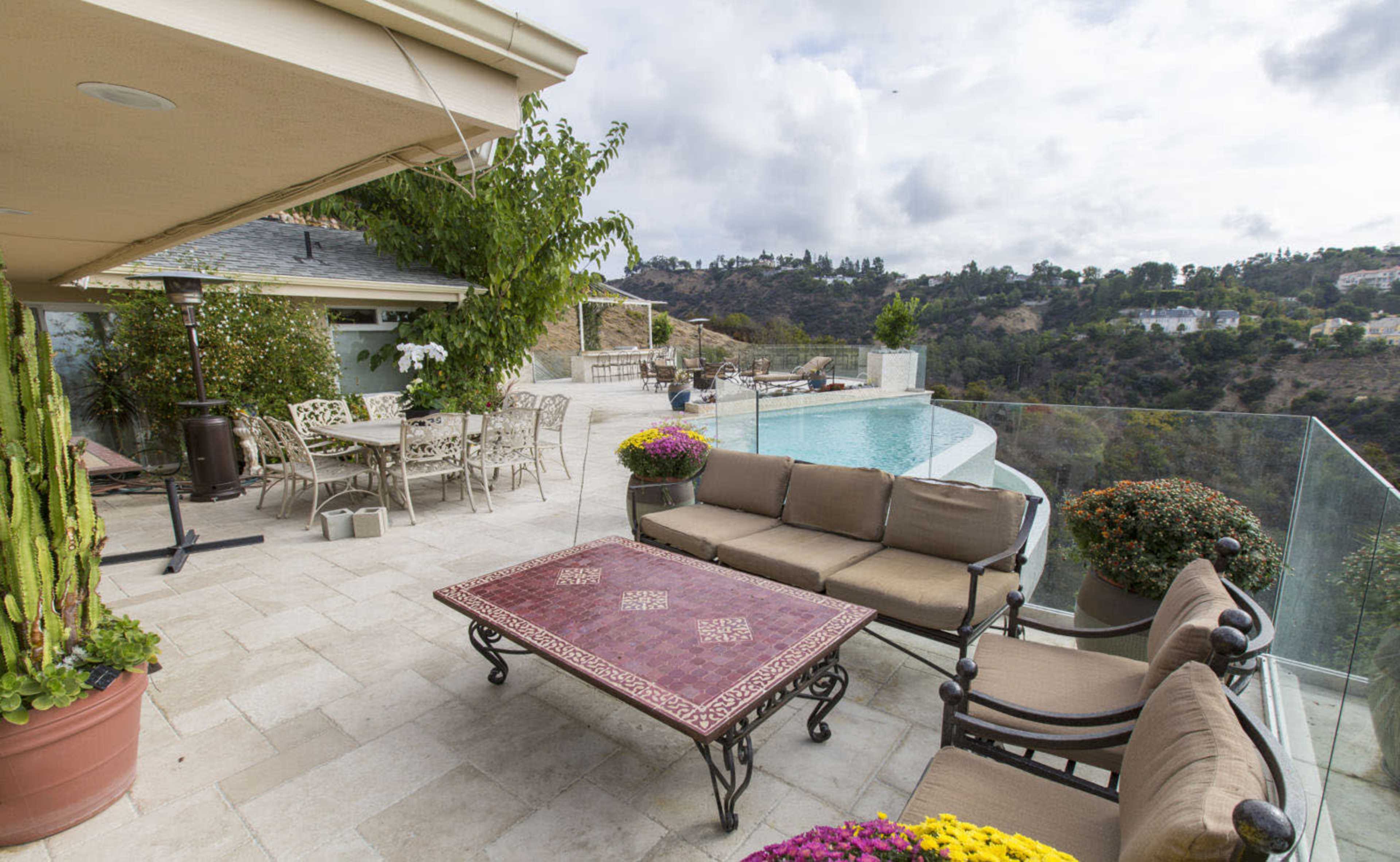 An outdoor patio features a seating area with a table, surrounded by potted plants and a view of a pool and hillside.