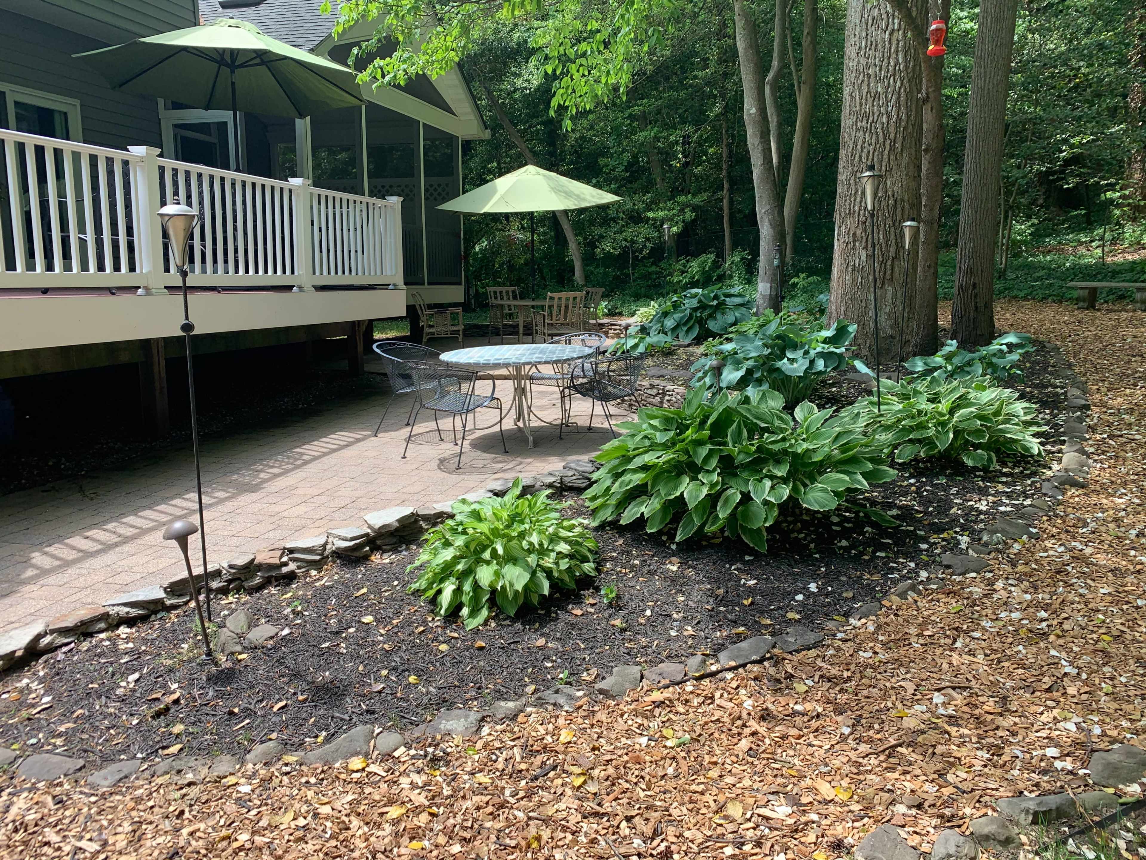 A circular stone patio with a table and chairs is surrounded by lush greenery and garden lights in a wooded area.