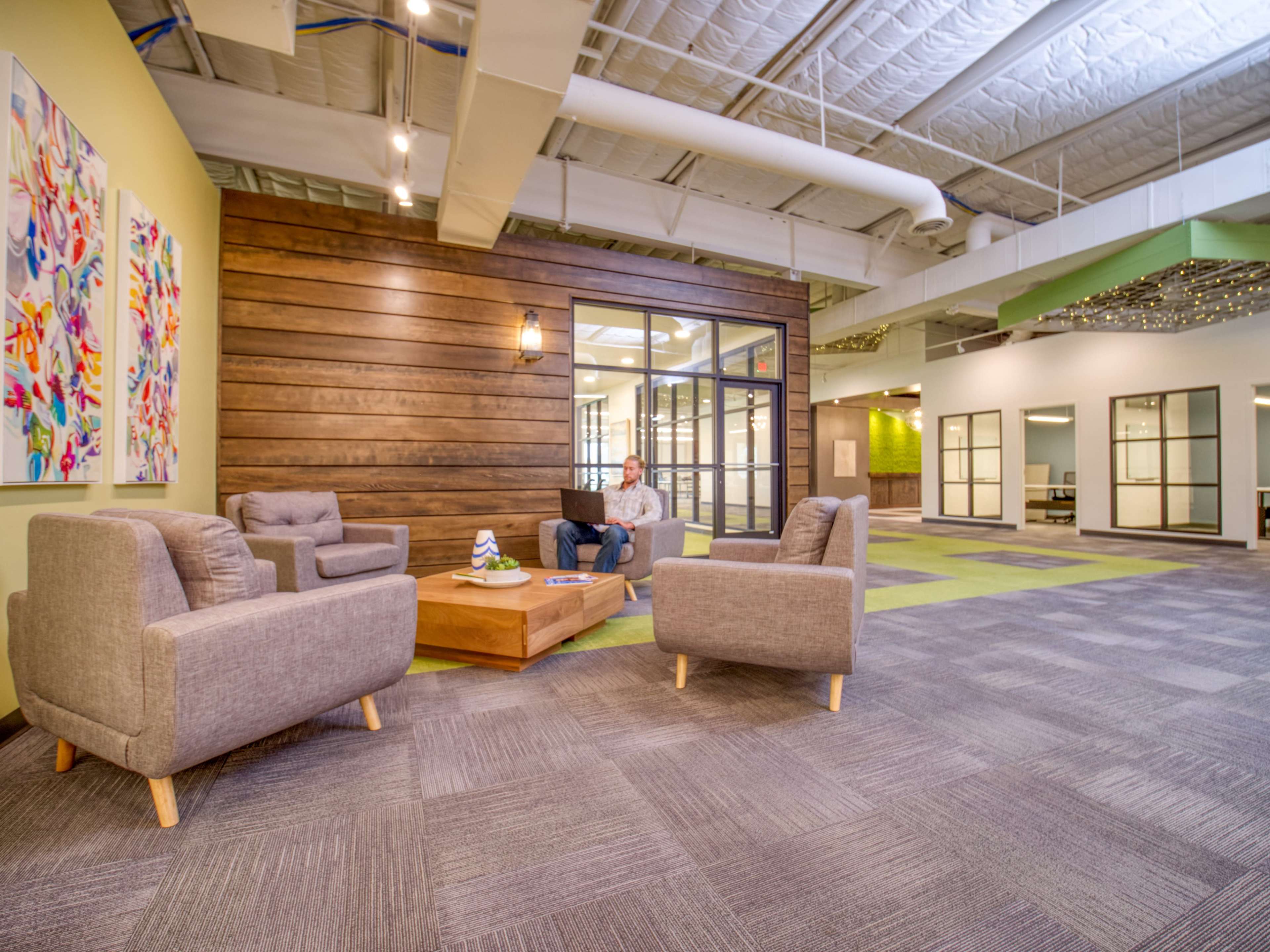 A modern office space features a seating area with gray chairs around a wooden coffee table, complemented by a colorful artwork on the wall and large windows.