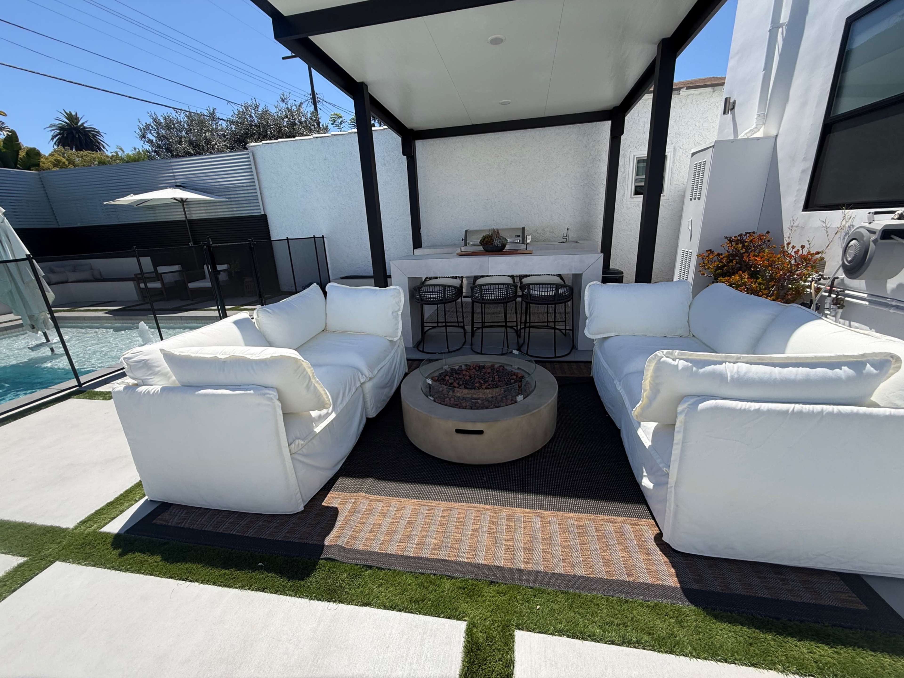 The outdoor seating area features two white sofas facing a central fire pit, with a bar and stools visible in the background beside a pool.