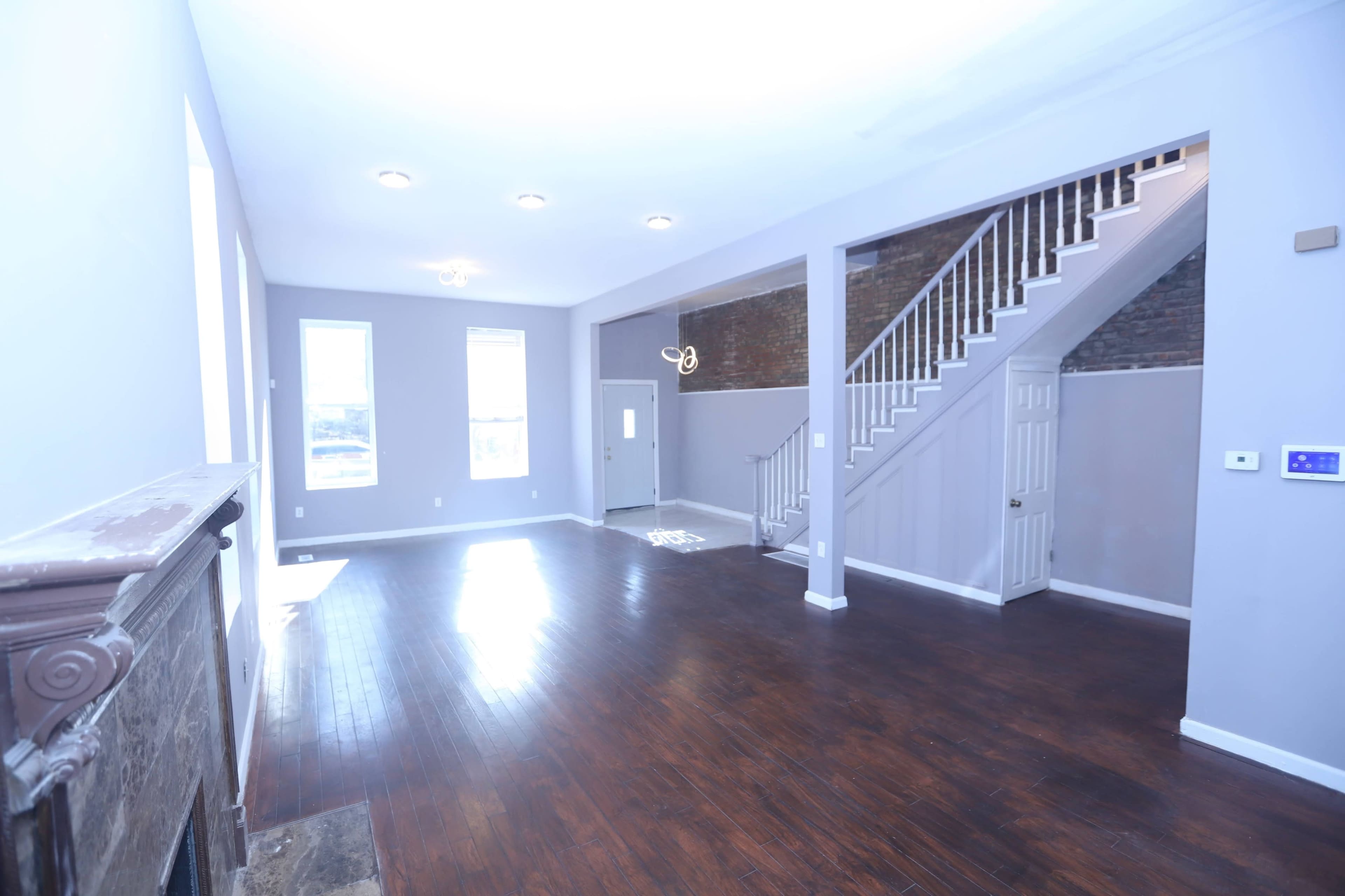 The image shows a spacious living room with hardwood floors, featuring large windows, a staircase, and a plain, light-colored wall.