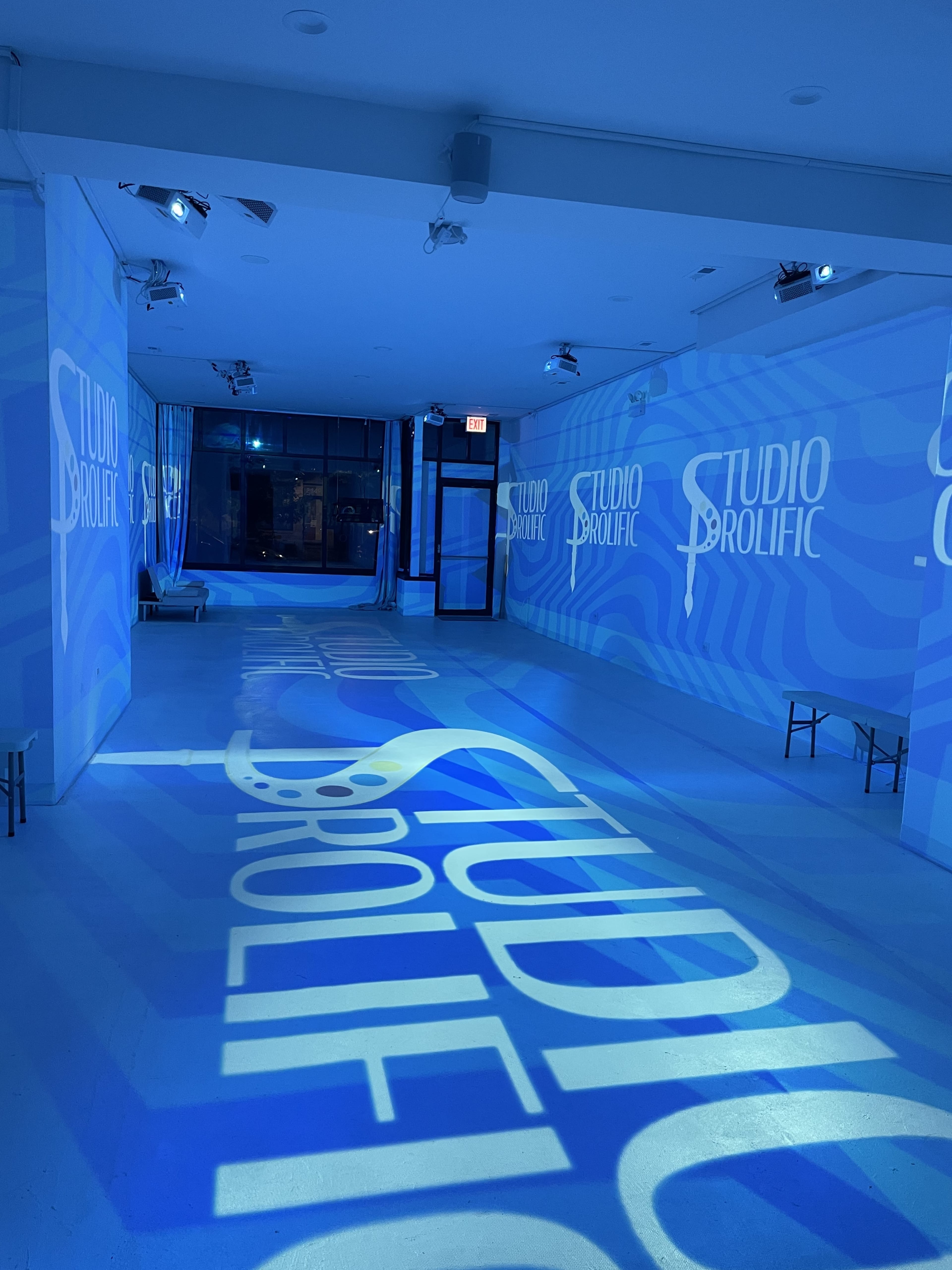 A spacious room with blue lighting and the words "STUDIO PROLIFIC" projected on the walls and floor.