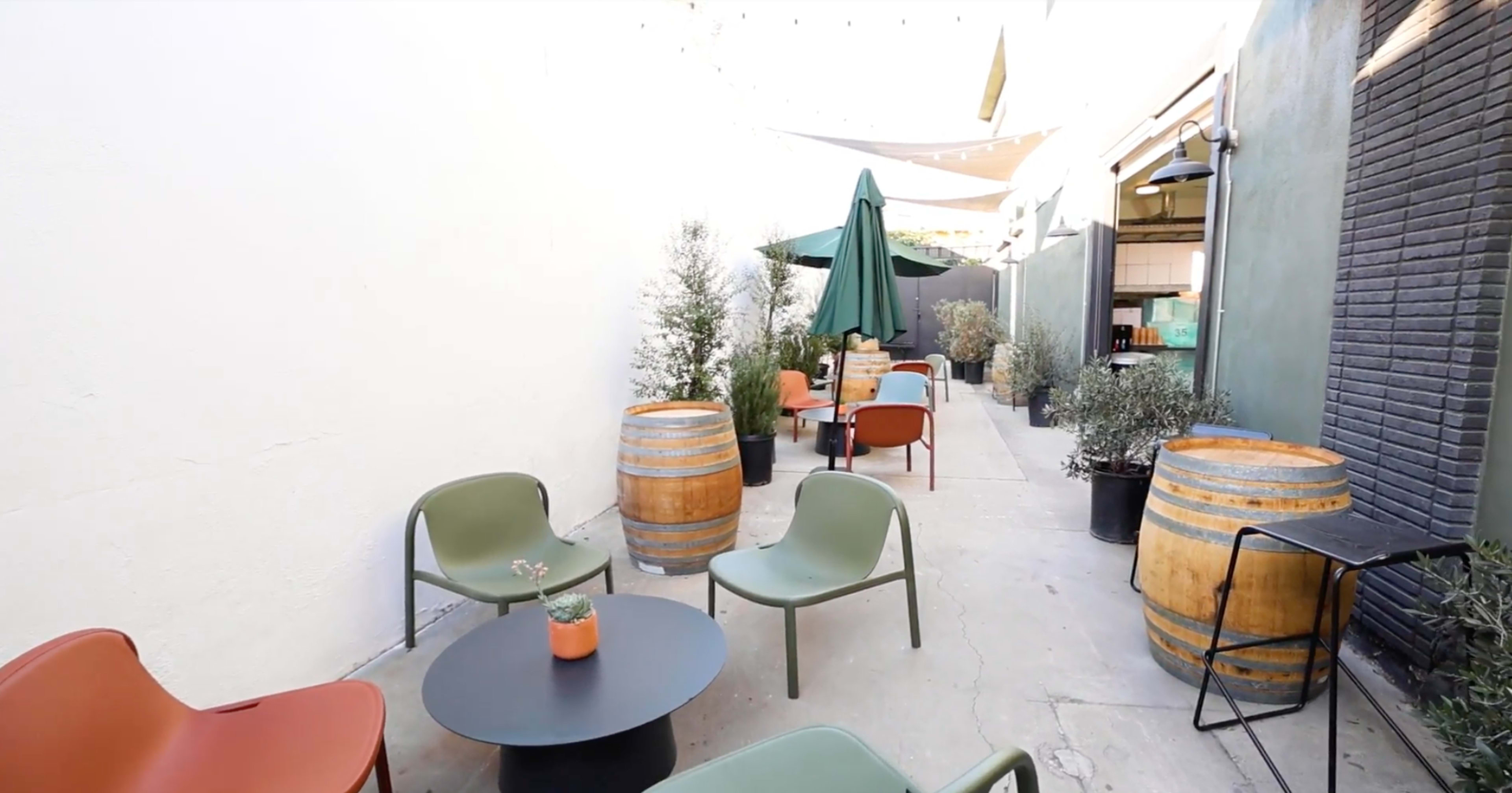 The image shows a narrow outdoor seating area featuring green and orange chairs around a black table, with wooden barrels and potted plants along the walls.