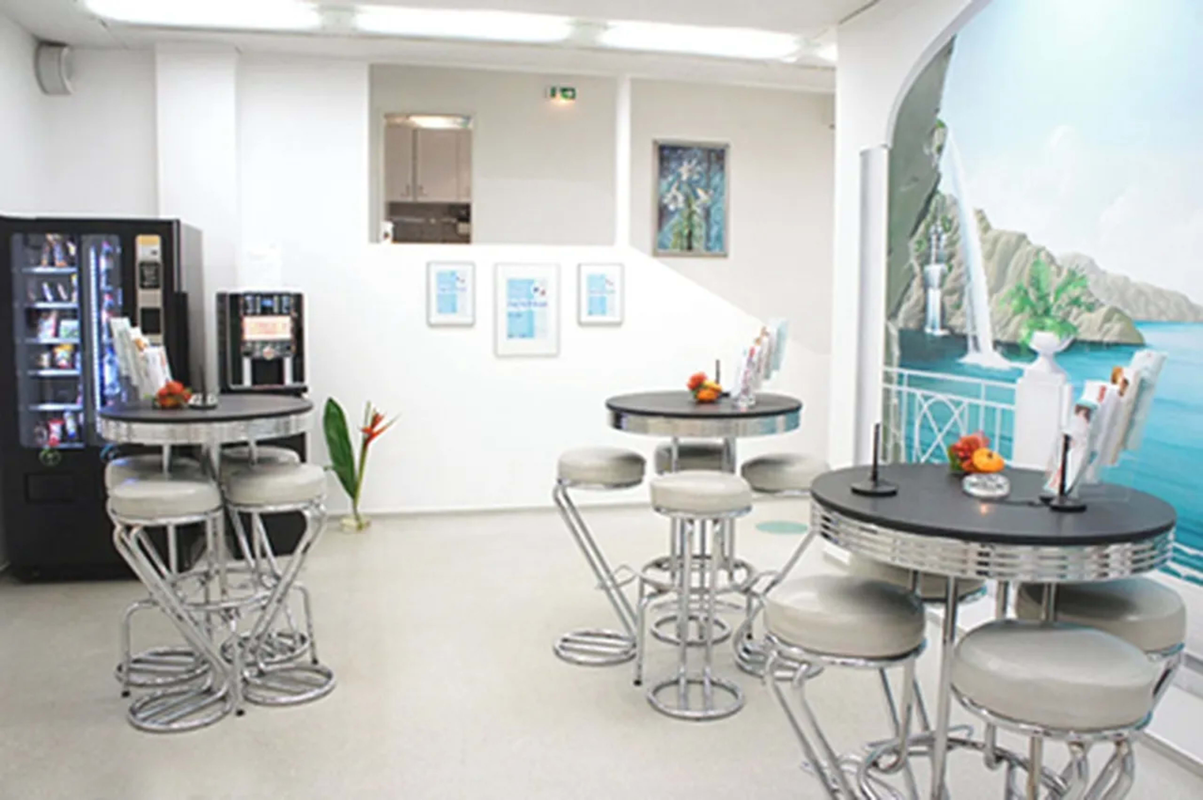 A bright, modern café featuring several round tables with stools, a vending machine, and a mural depicting a coastal scene on the wall.