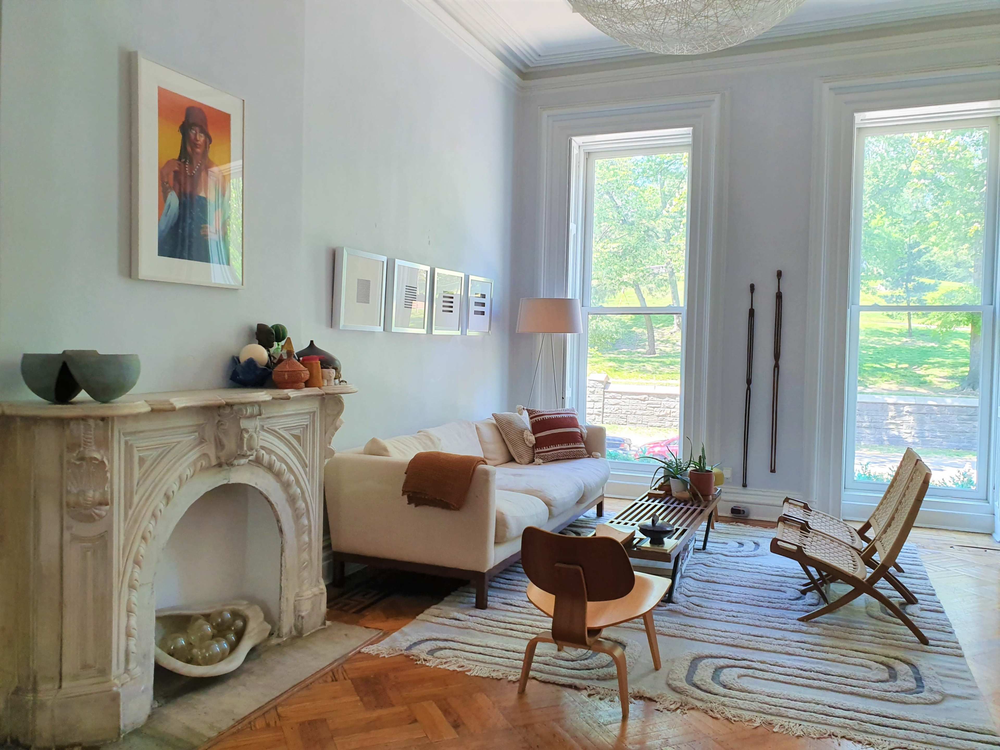 A bright living room featuring a white sofa, an ornate fireplace, and large windows with a view of greenery outside.