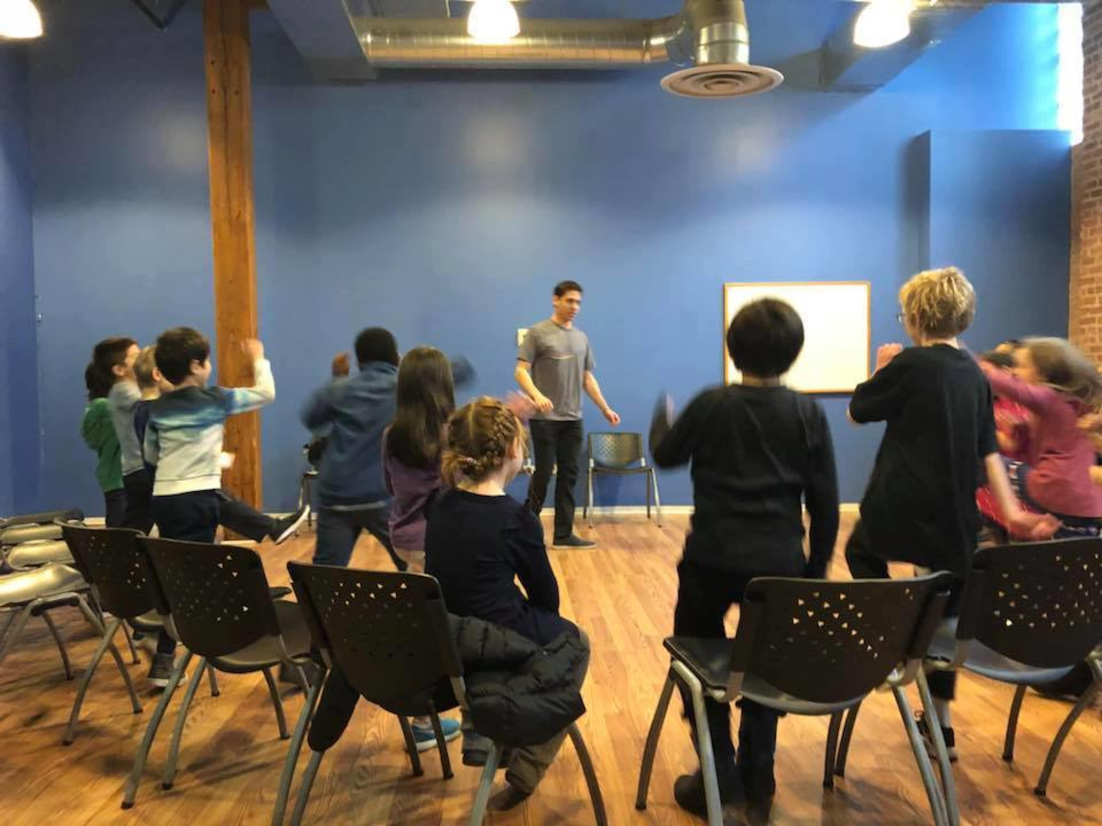 A group of children participates in a lively activity while seated in chairs facing an adult in a blue room.