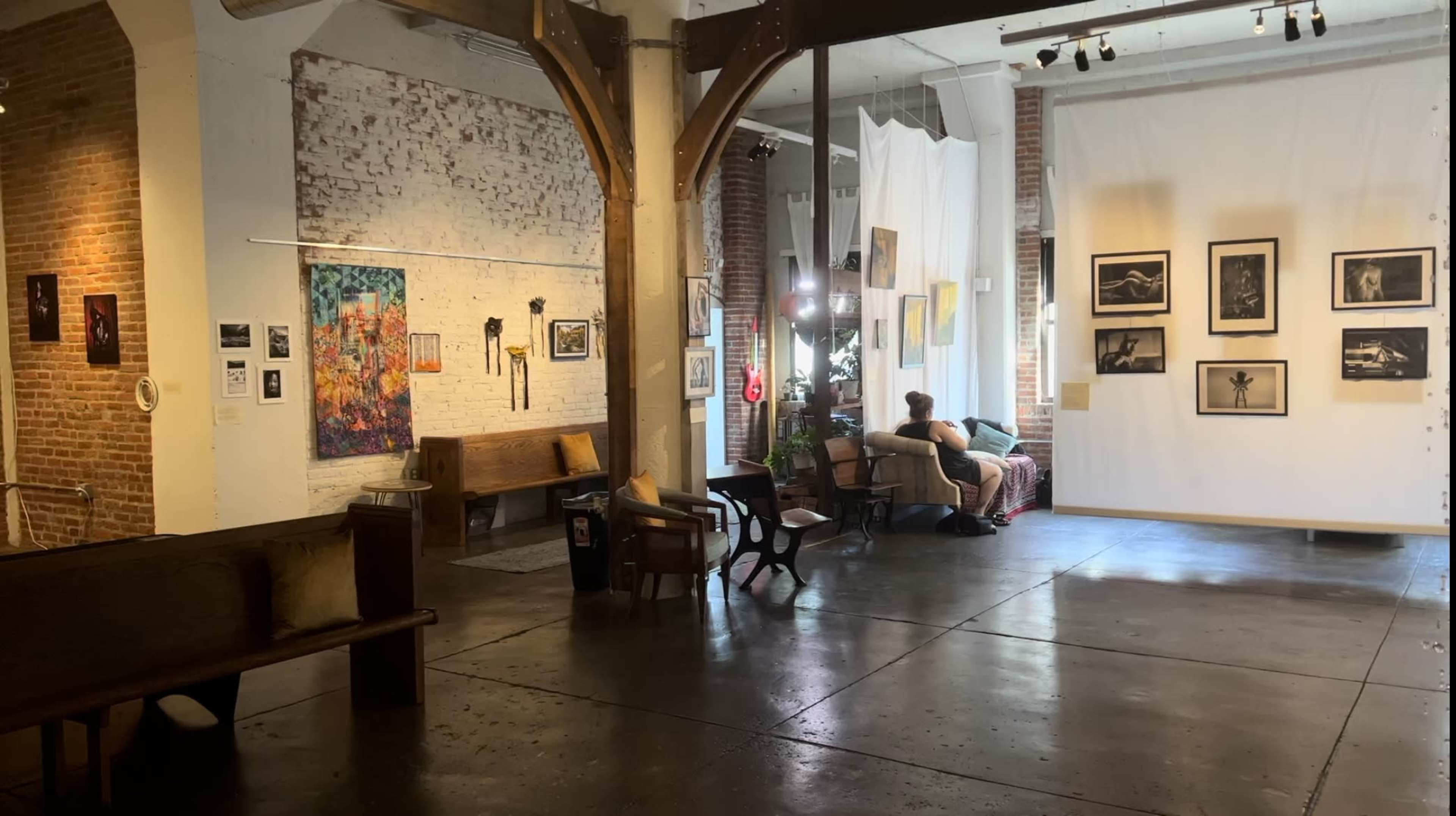 An art gallery with exposed brick walls, wooden beams, and various artworks displayed, alongside seating arrangements and a person sitting on a couch.