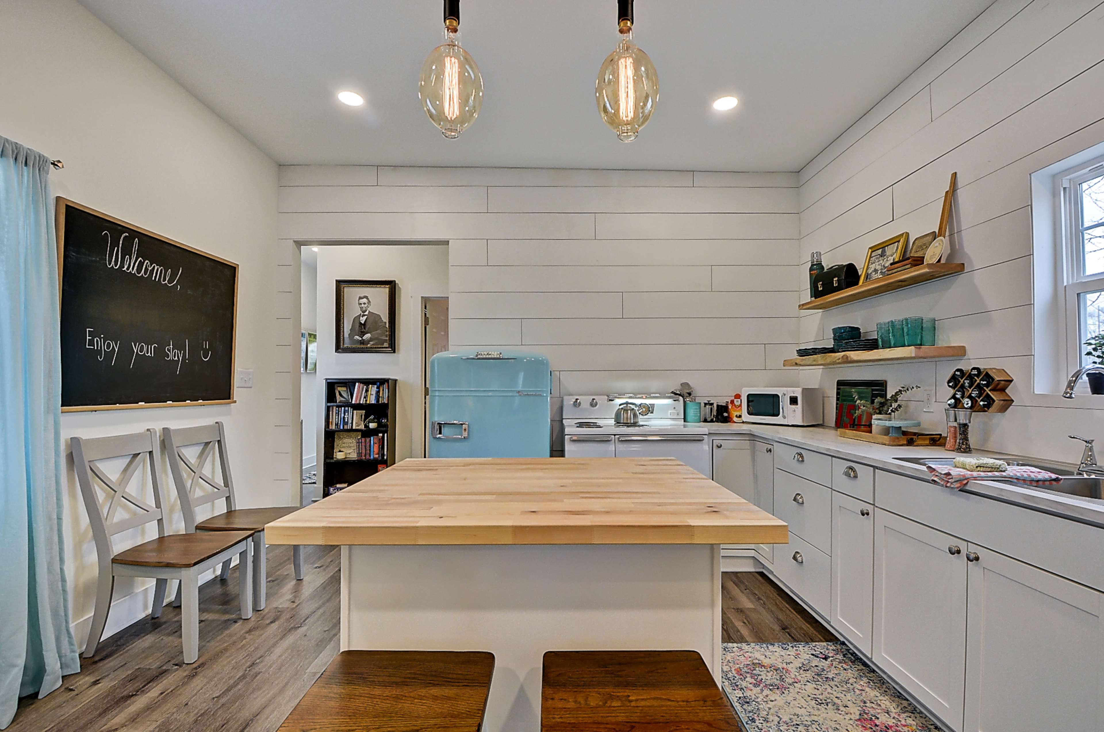 A modern kitchen featuring a large wooden island, white cabinets, a vintage style blue refrigerator, and a chalkboard welcoming guests.