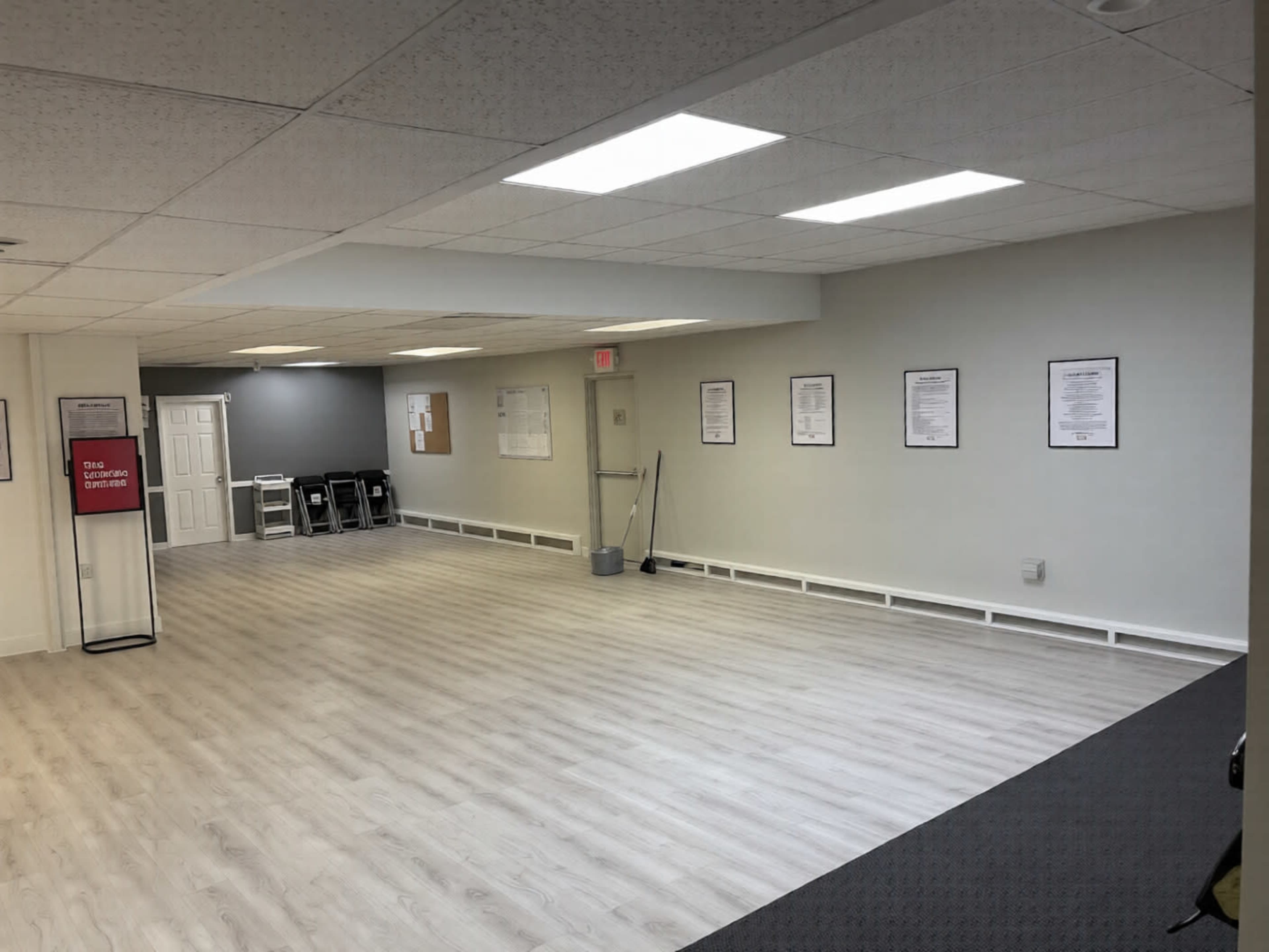 The image shows a spacious, empty room with gray walls and a light-colored floor, featuring several framed documents on the walls and chairs arranged against one side.