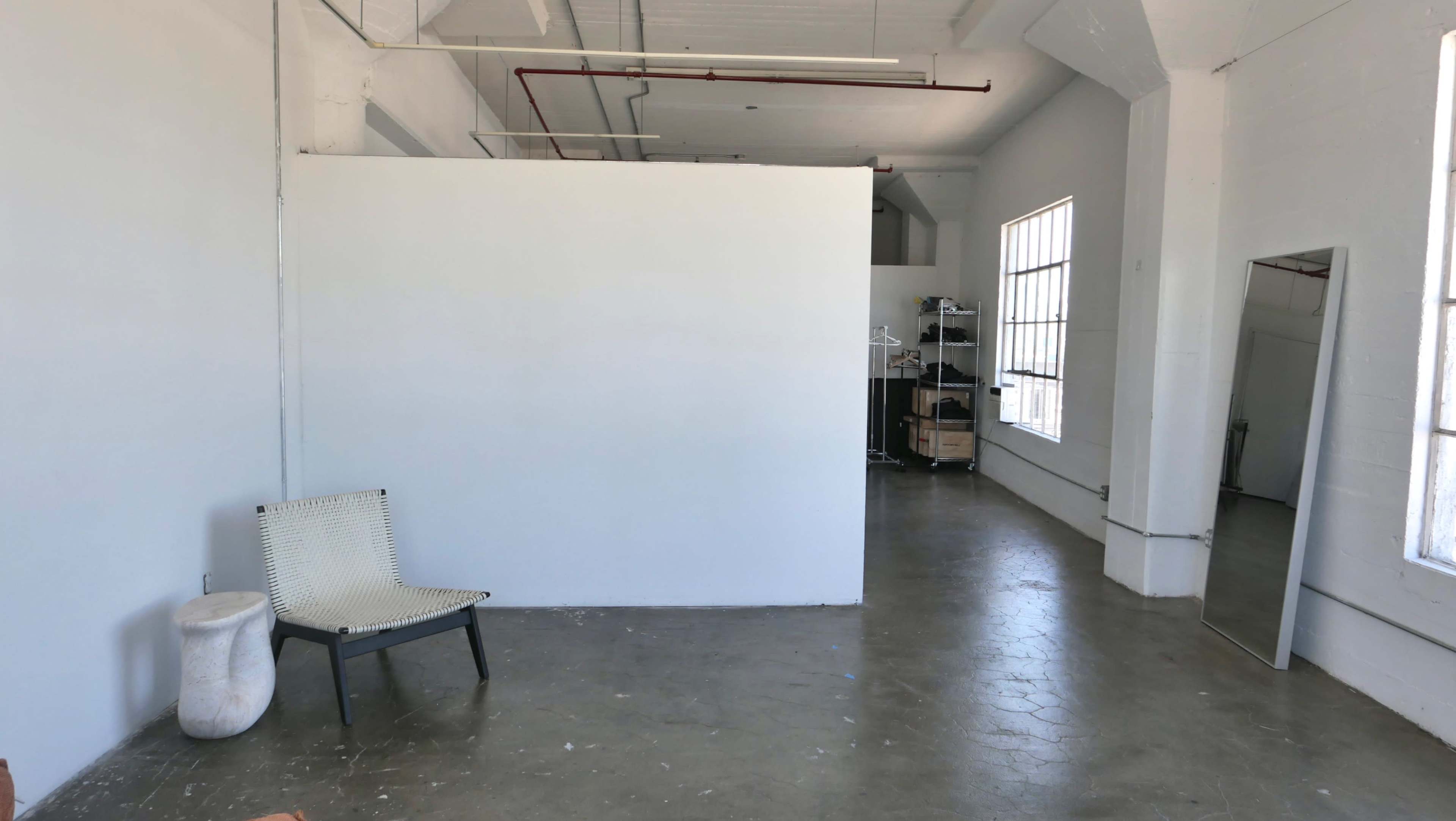 An empty room features a white wall, a chair, a large mirror, and a shelving unit in the background.