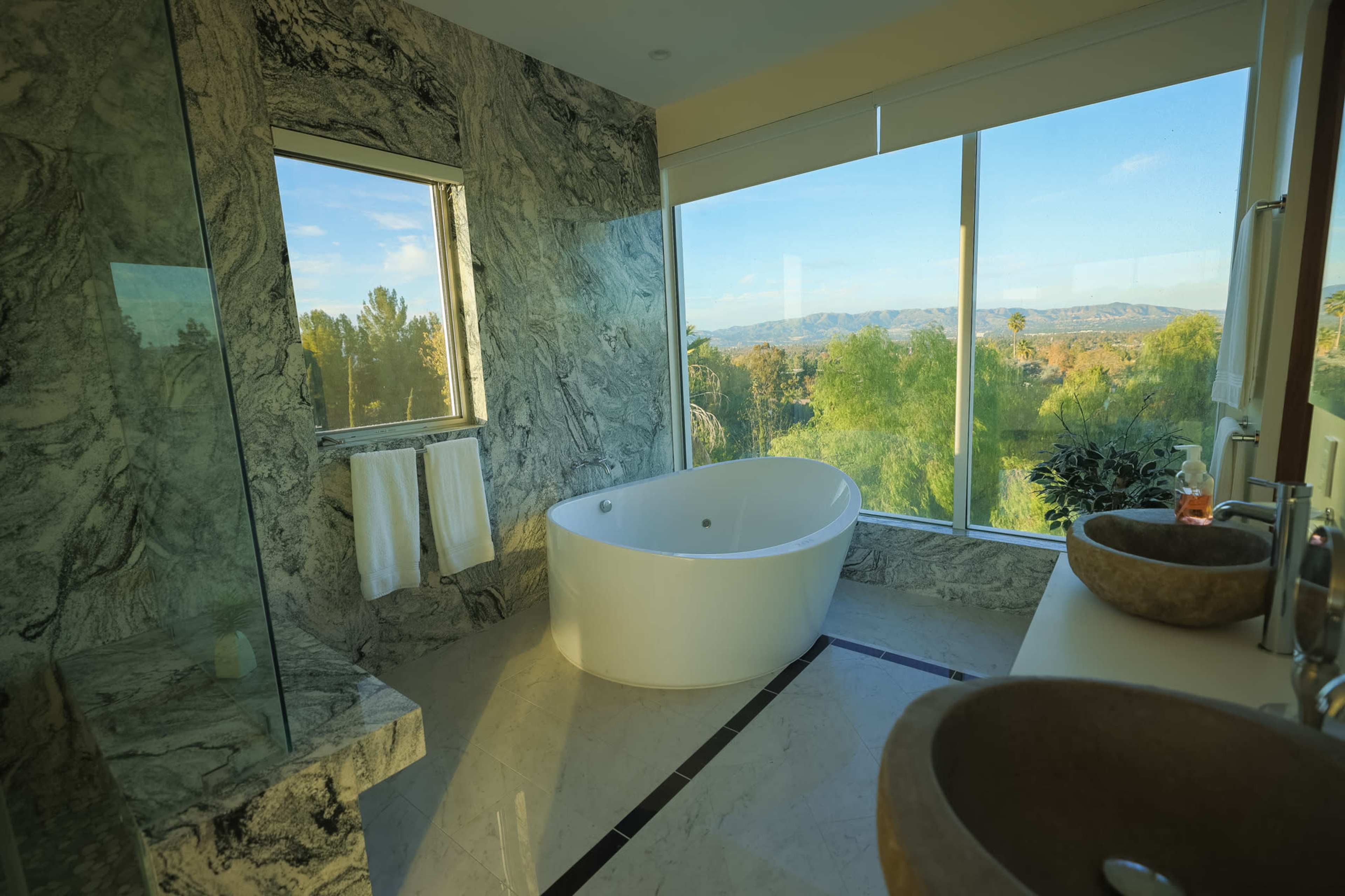 The bathroom features a freestanding white tub, two stone sinks, and large windows offering a view of the surrounding landscape.