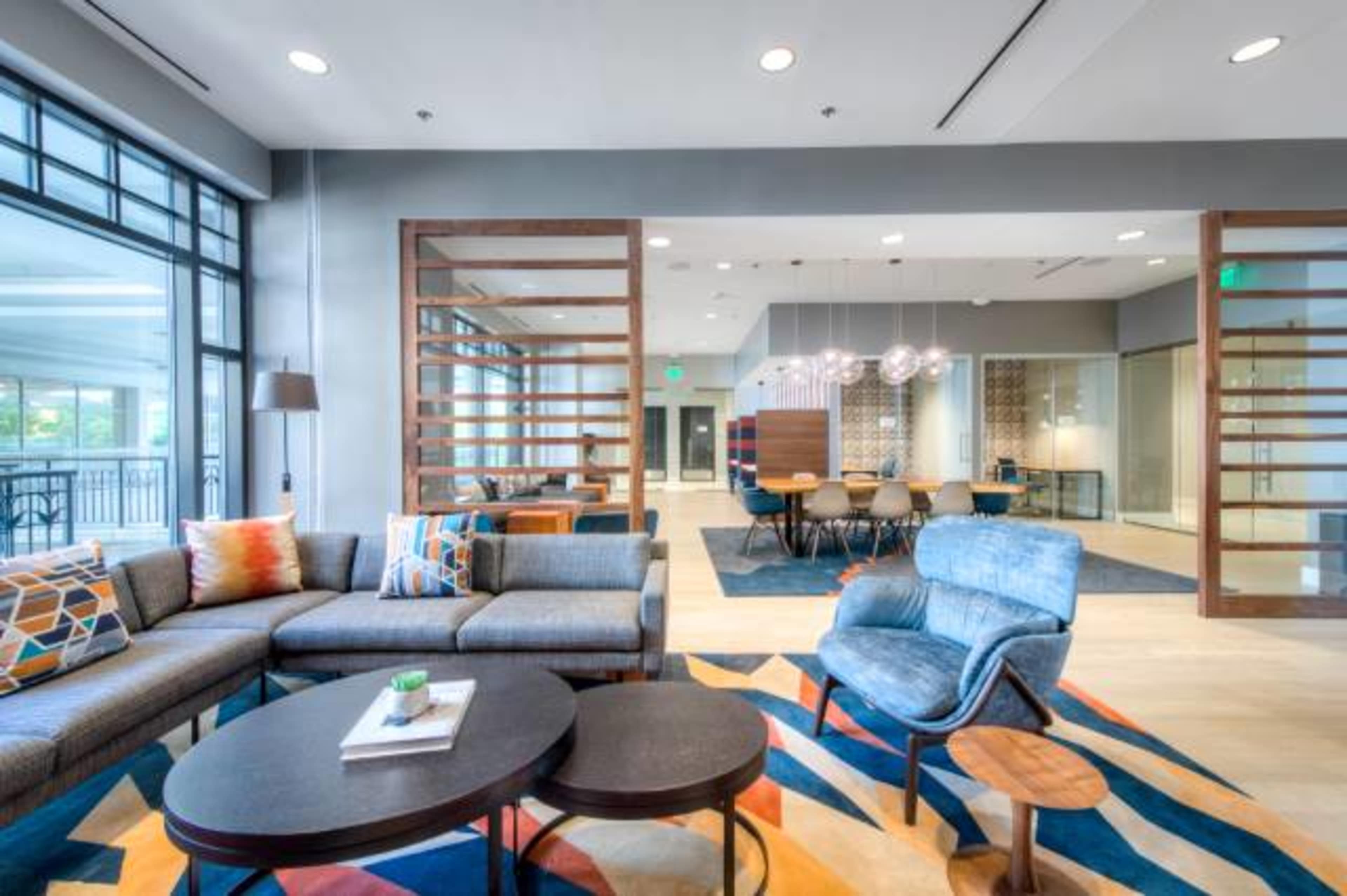 A modern lounge area with a sectional sofa, coffee tables, and a dining space featuring a large table and chairs, all arranged in a spacious, brightly lit room.
