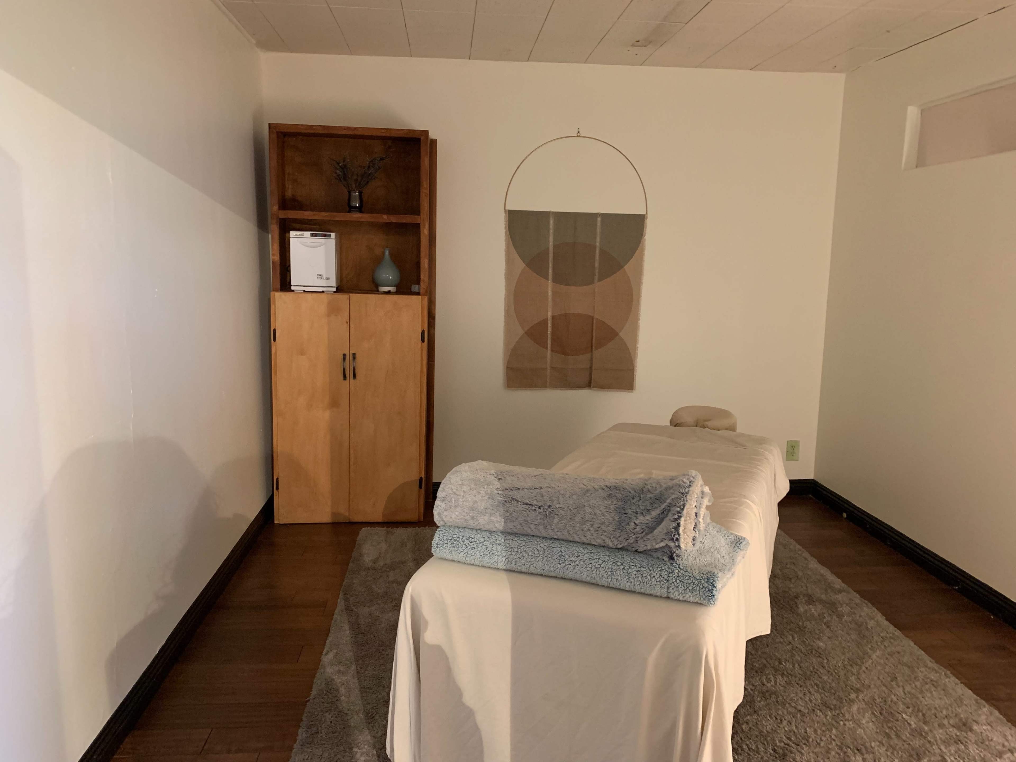 The image shows a massage therapy room with a treatment table, neatly arranged towels, and wooden storage unit against a white wall.
