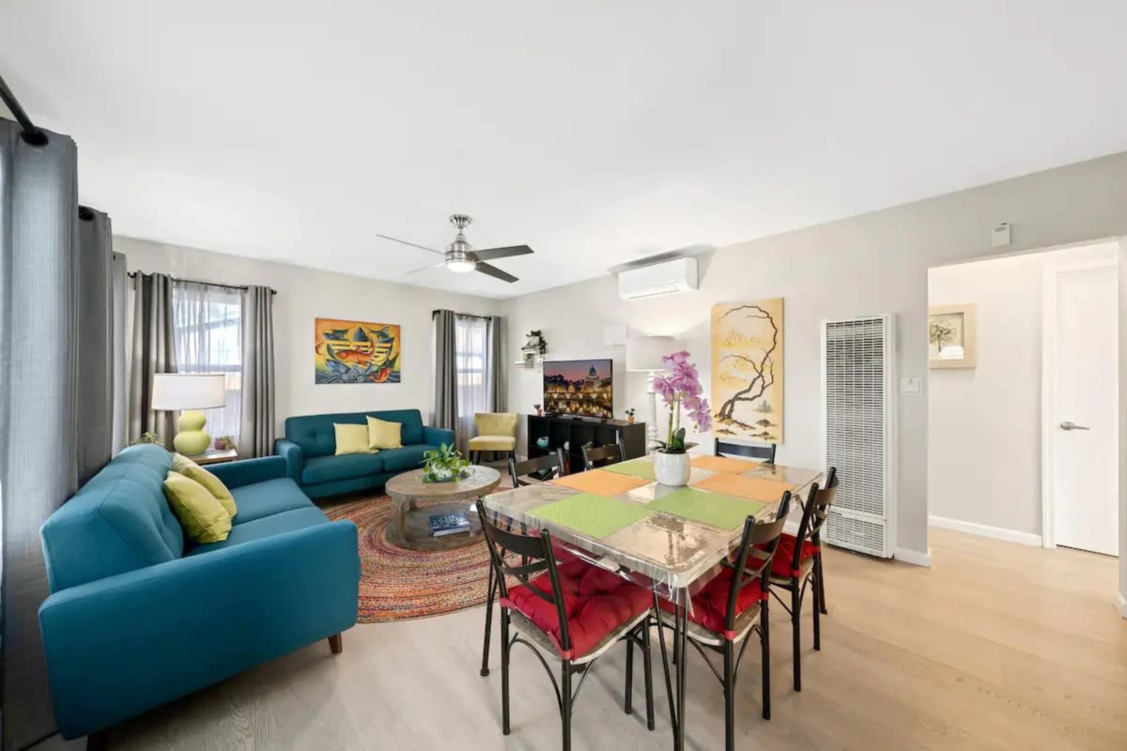 A brightly lit living space features a teal sofa and a glass dining table surrounded by chairs, with decorative artwork on the walls.