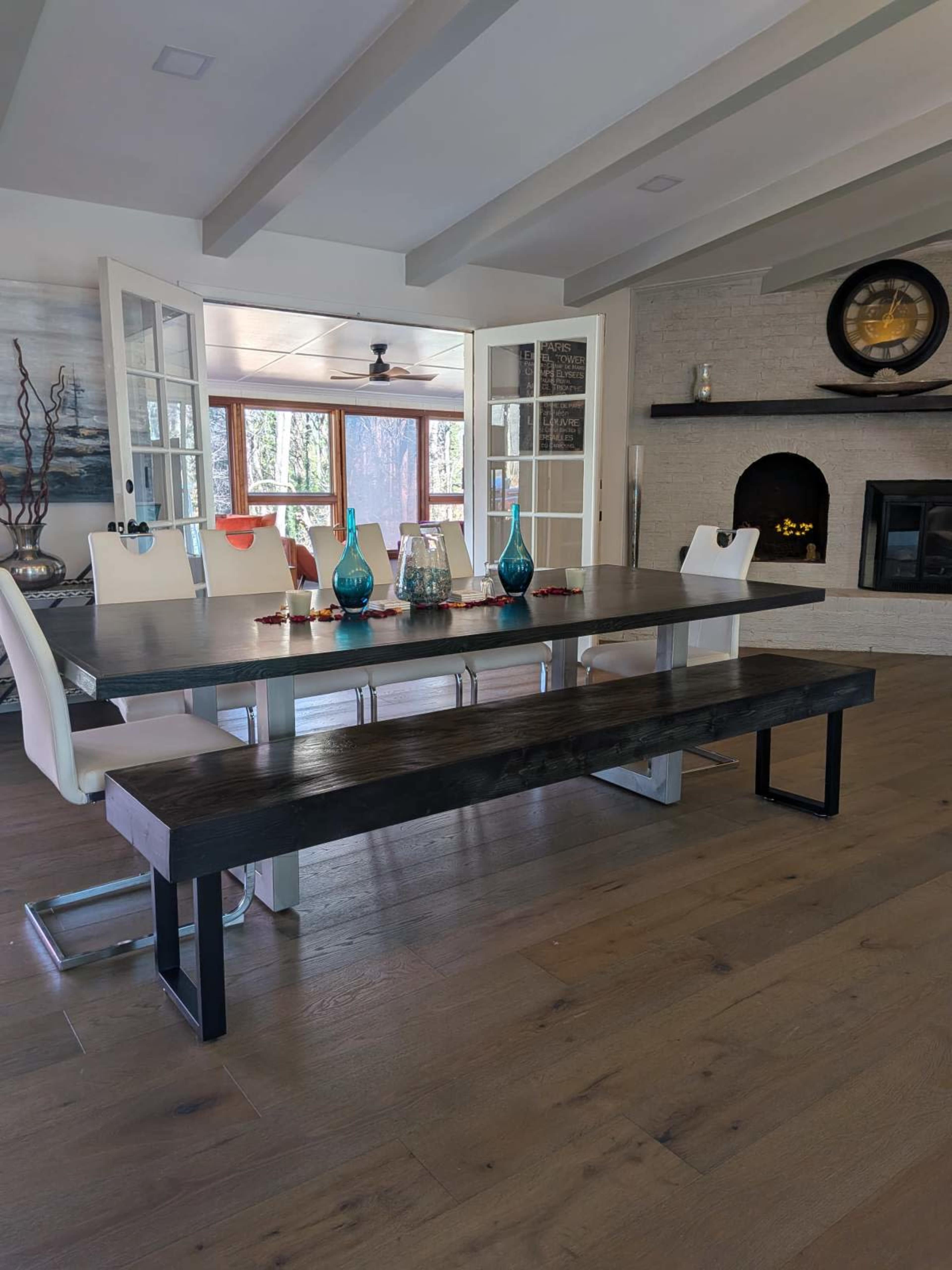 A modern dining area features a large wooden table with seating for six, flanked by a long bench, and large glass doors that open to a sunlit room.