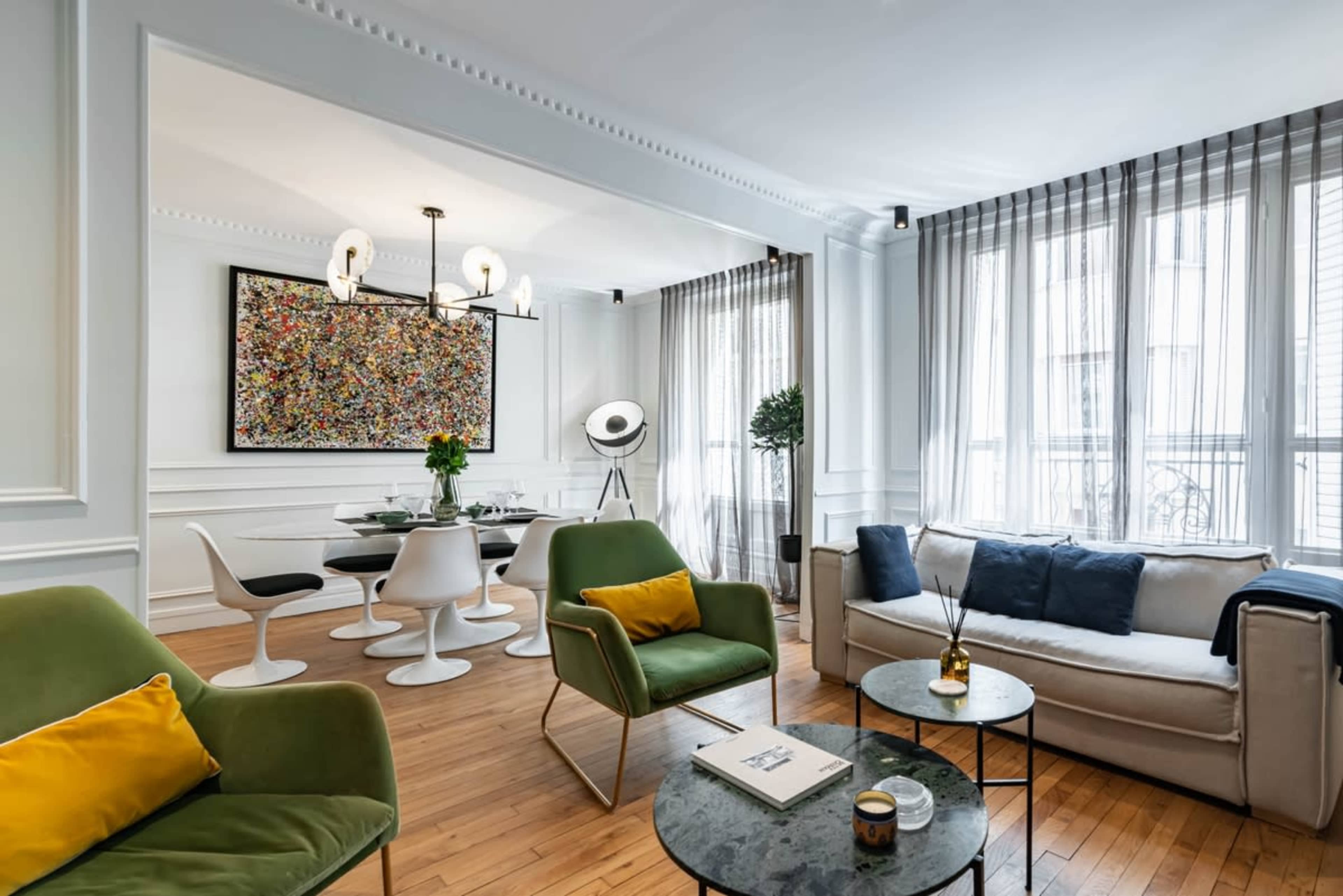 A modern living room with a mix of green and white furniture, a round table with white chairs, and large windows draped with sheer curtains.