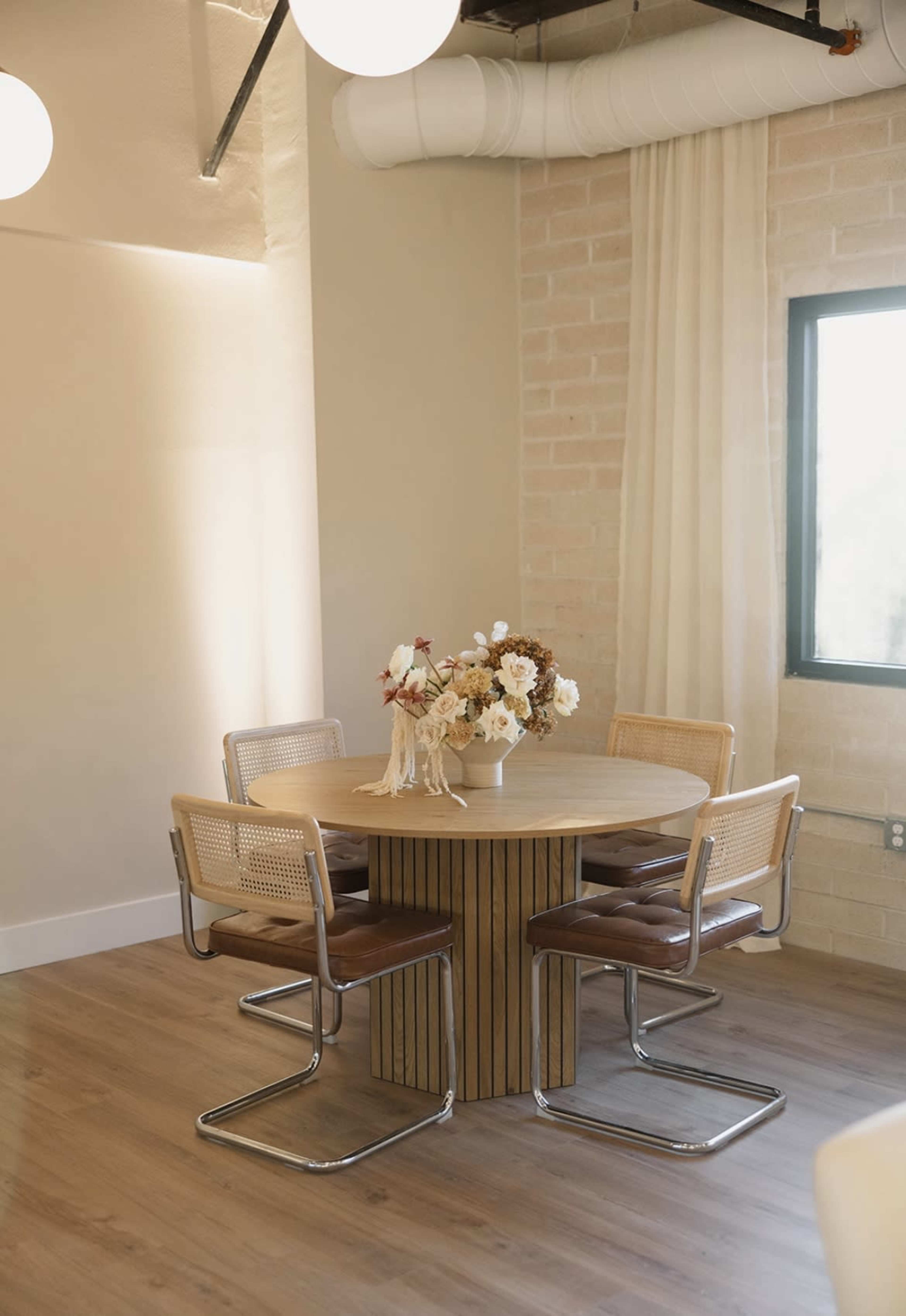 A round wooden table with a vertical slatted base is surrounded by four modern chairs featuring cane backs and brown leather seats in a well-lit room.