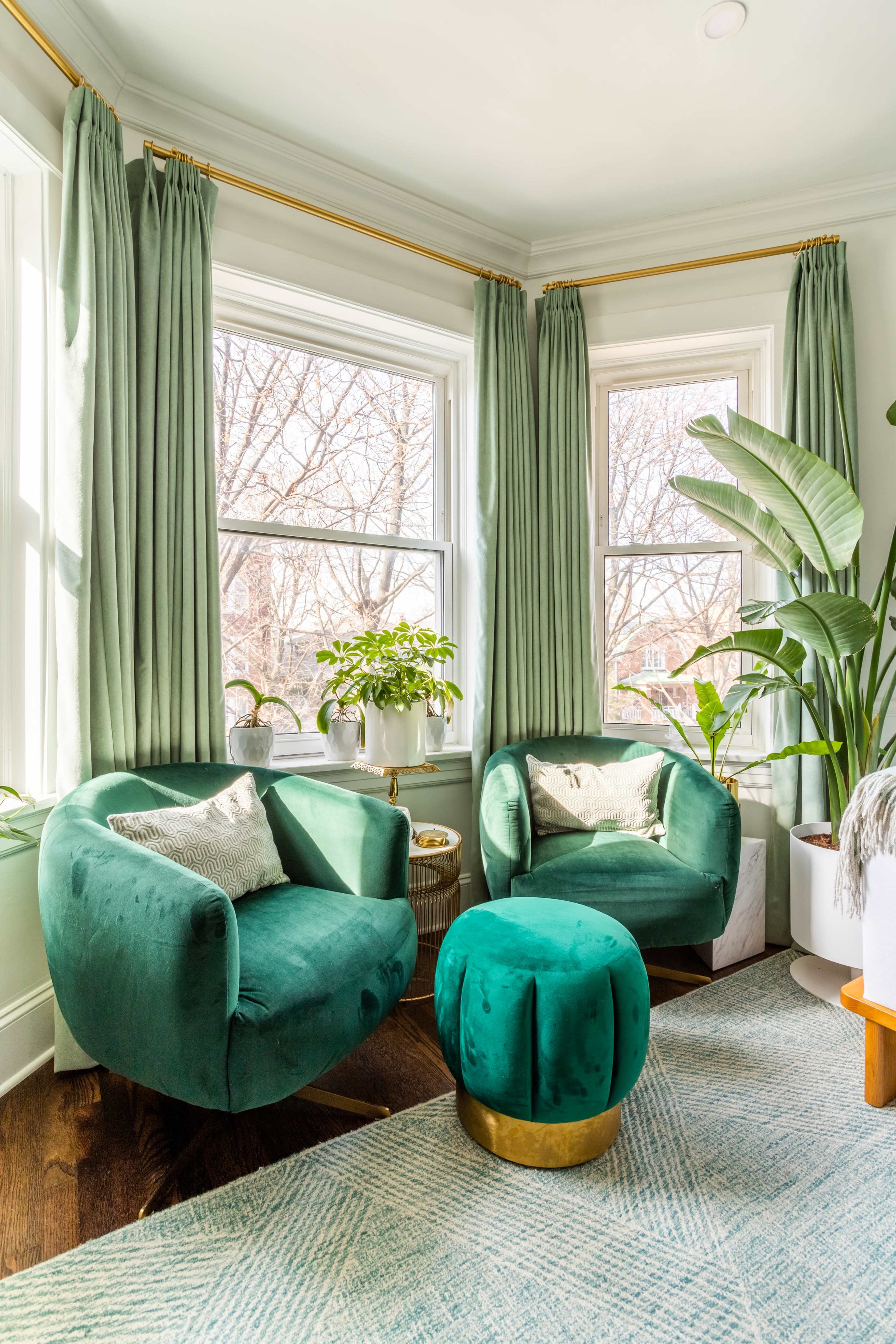 A bright corner of a living room featuring two green velvet armchairs, a round green ottoman, potted plants, and large windows with sheer curtains.