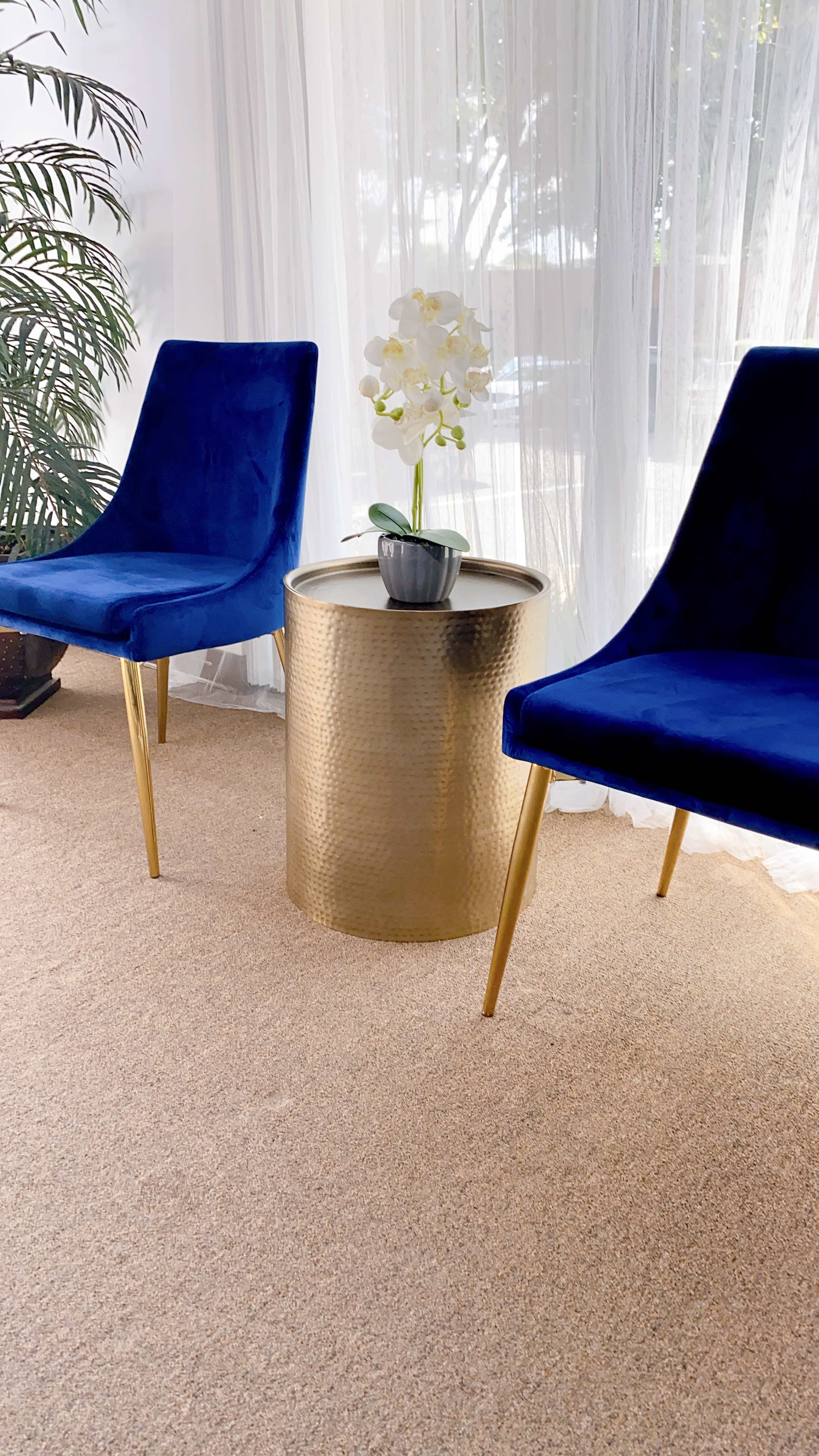 Two blue chairs flank a gold, round side table with a potted orchid, set against a backdrop of sheer curtains and a nearby plant.
