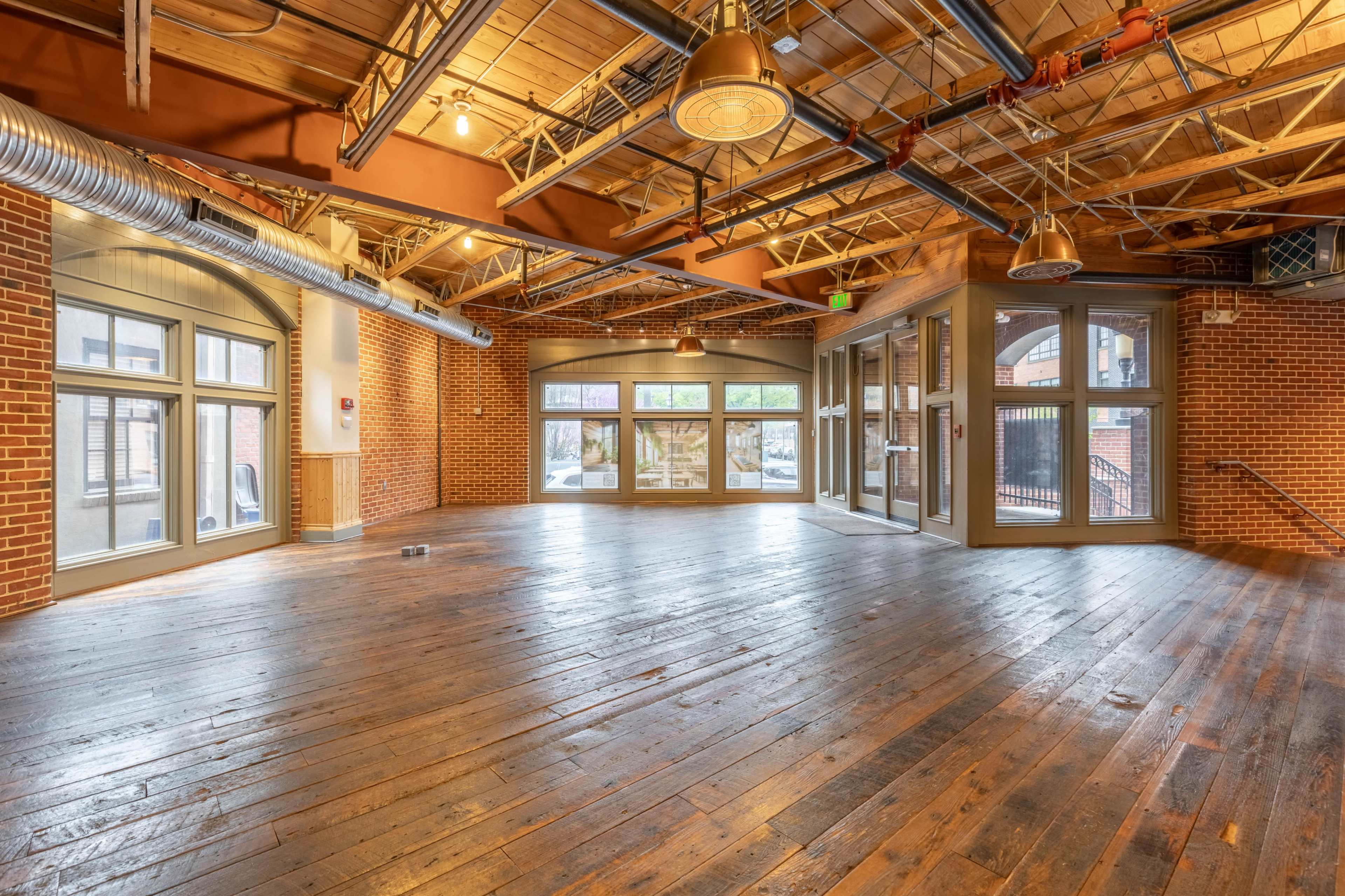 The image shows a spacious indoor area with wooden floors, exposed beams, and large windows that open to an outdoor terrace.