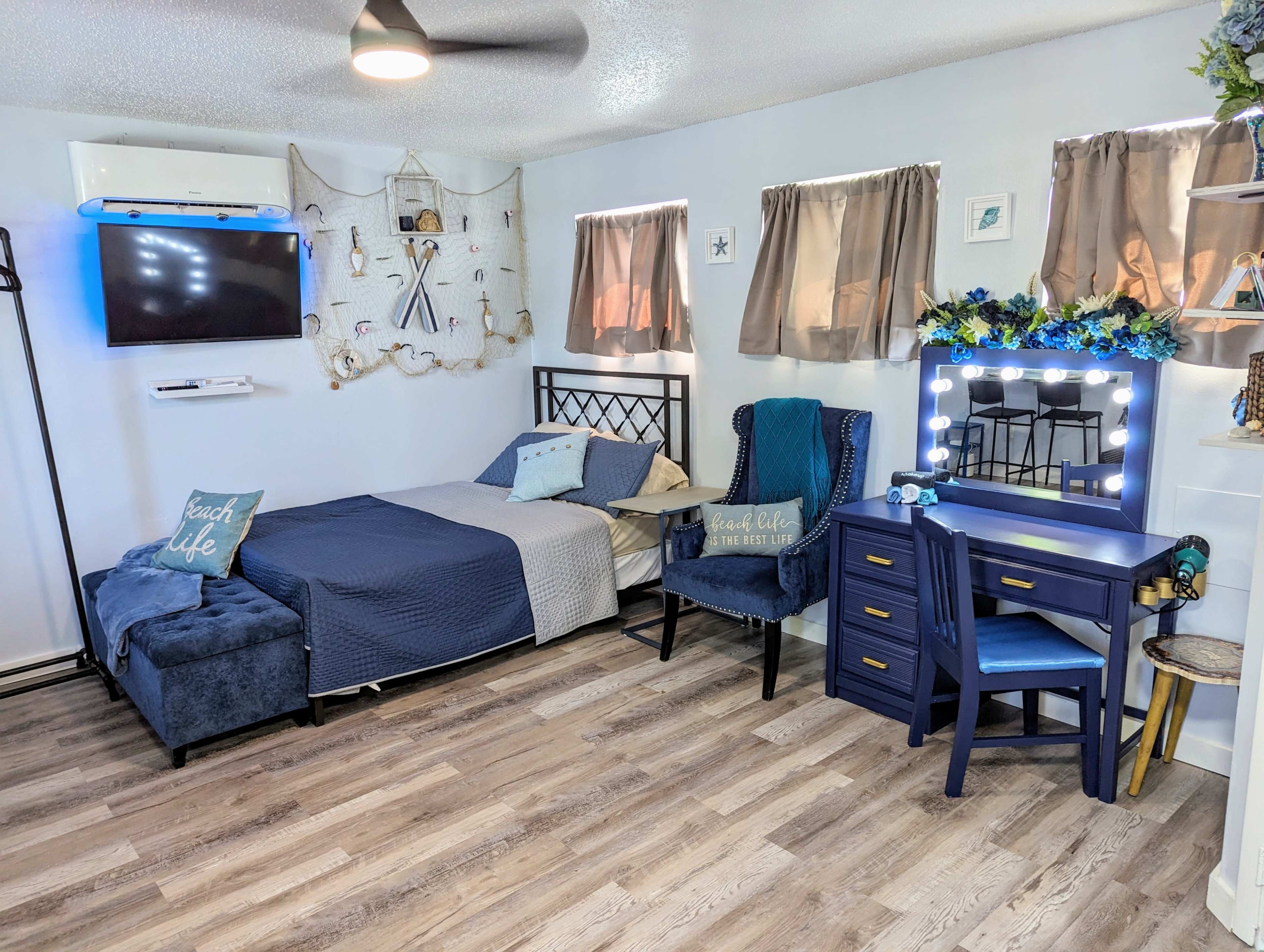 A neatly arranged bedroom featuring a bed with a blue cover, a matching chair and mirror, a television mounted on the wall, and decorative items on shelves.