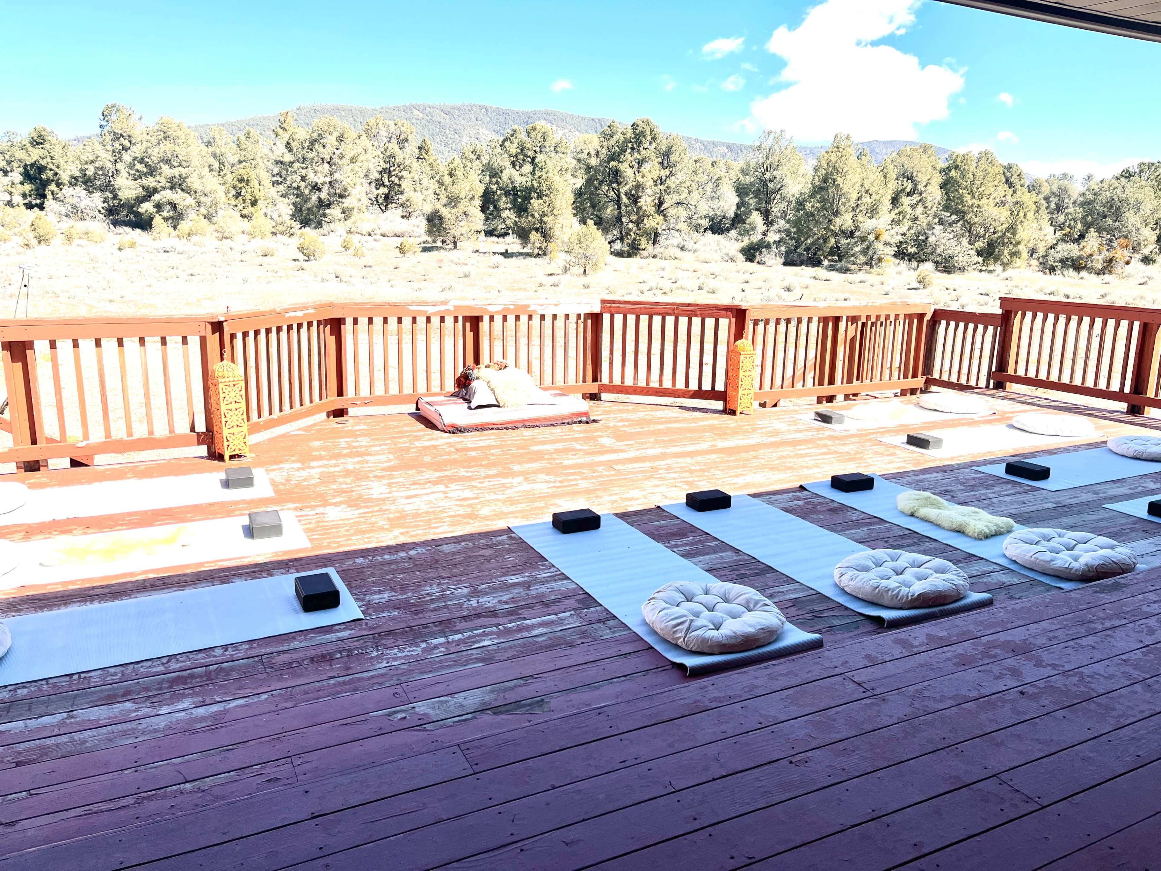 The image shows a spacious wooden deck with meditation cushions and mats arranged neatly, overlooking a landscape of trees and mountains.