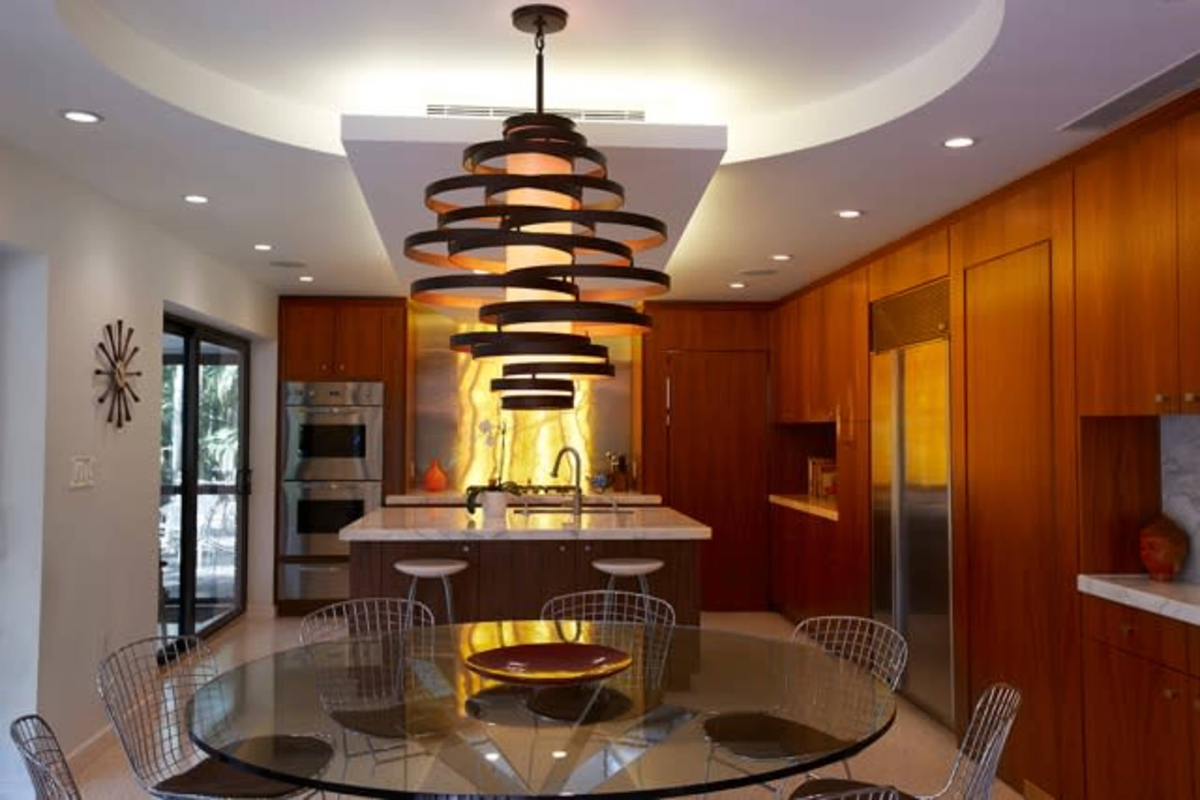 A modern kitchen features wooden cabinetry, a central island with a circular glass dining table, and a distinctive pendant light.