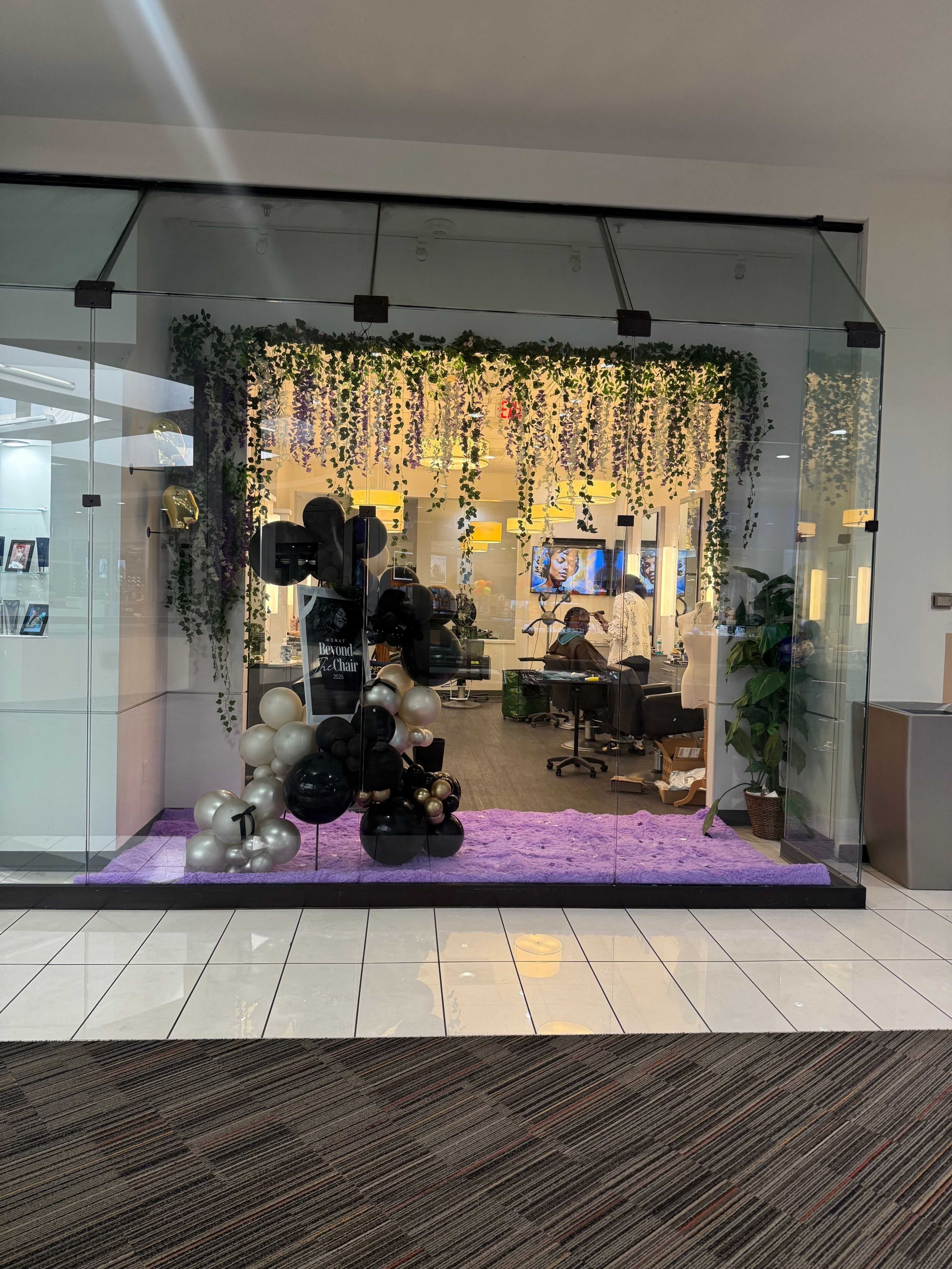 A glass storefront displays black, white, and gold balloons adorned with greenery, set against a purple carpet inside a modern space with bright lighting.
