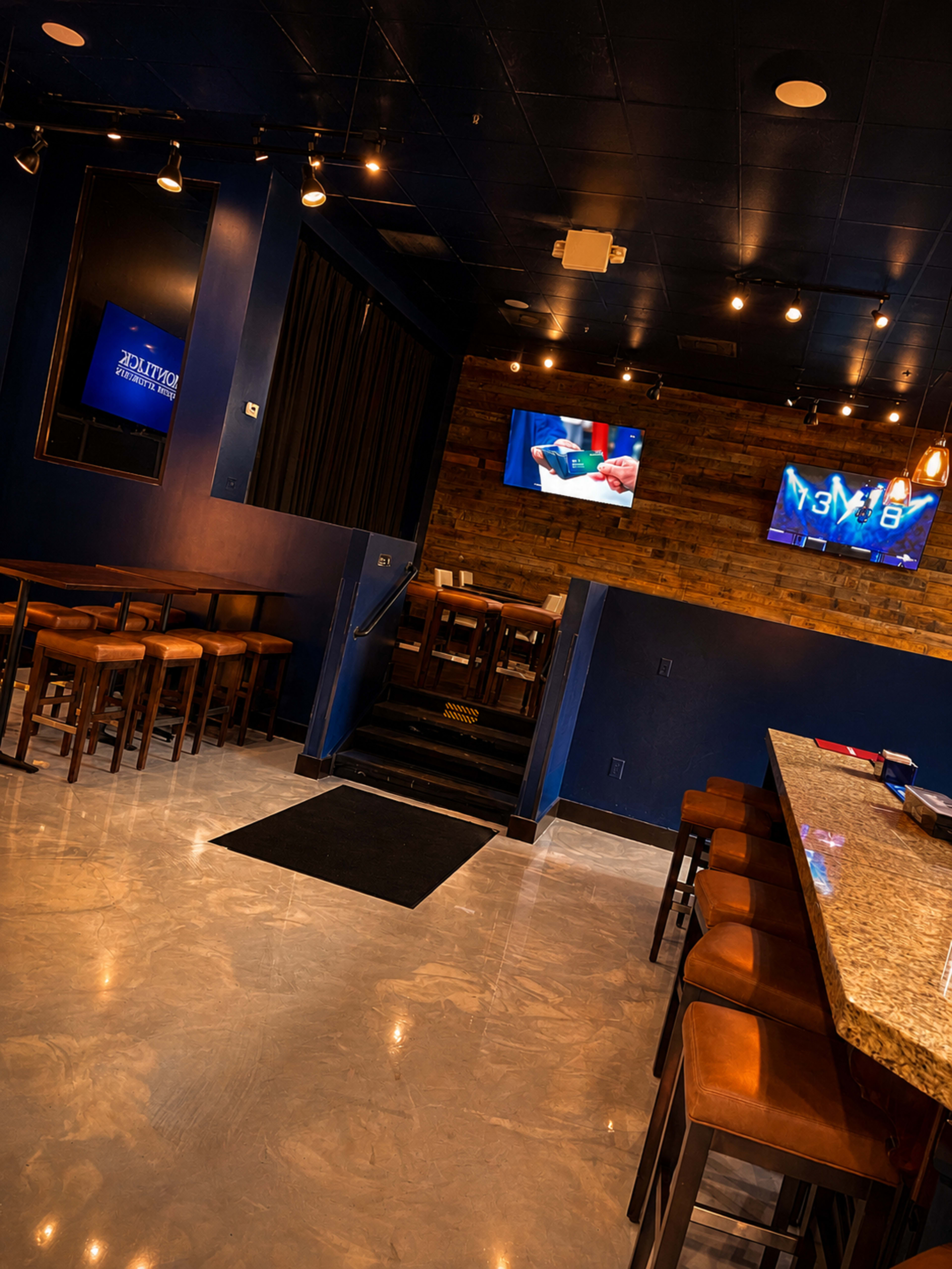 The interior of a modern bar features a long granite counter, wooden accents on the walls, and multiple TVs mounted on the dark-colored backdrop.
