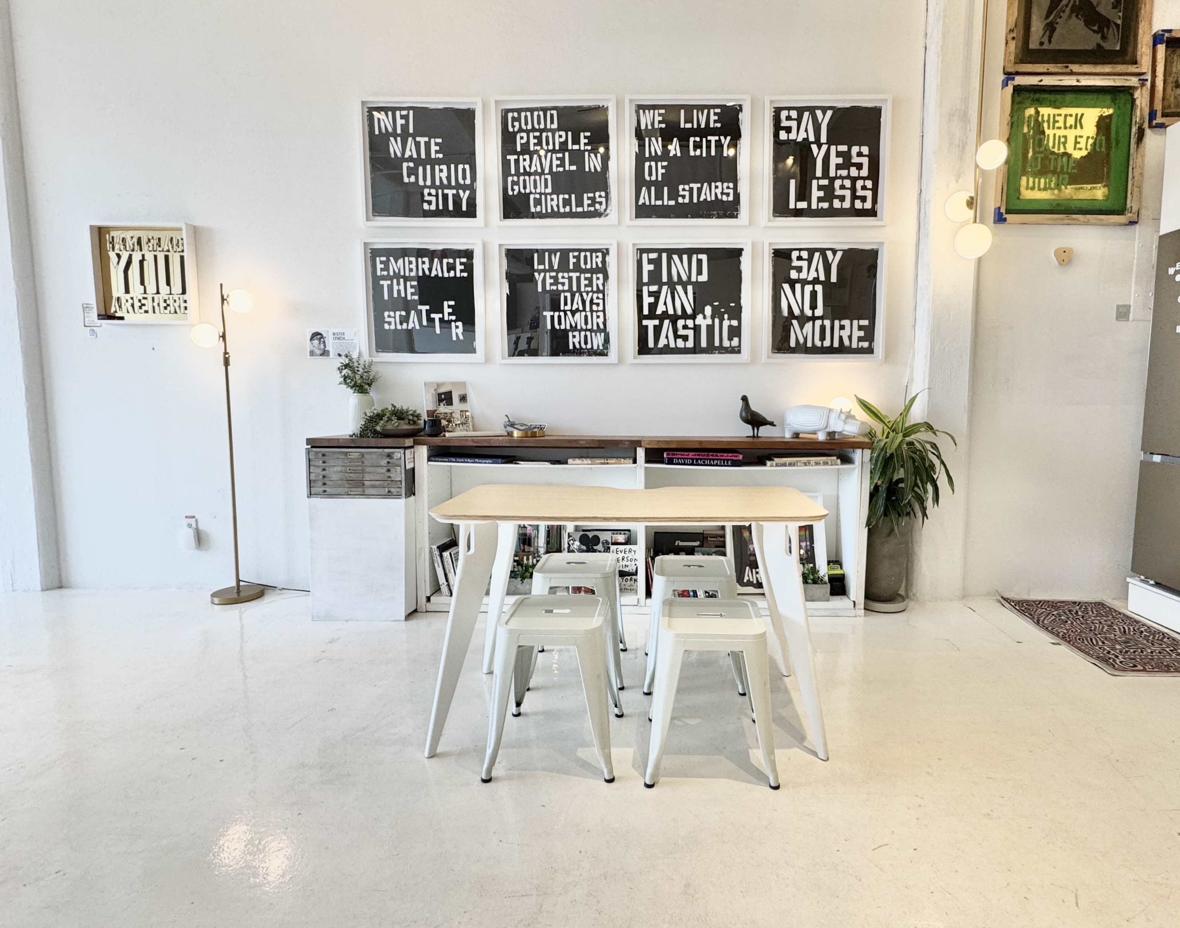 The image shows a minimalist interior design featuring a table and two chairs, with a wall display of framed black-and-white signs and a stylish floor lamp.