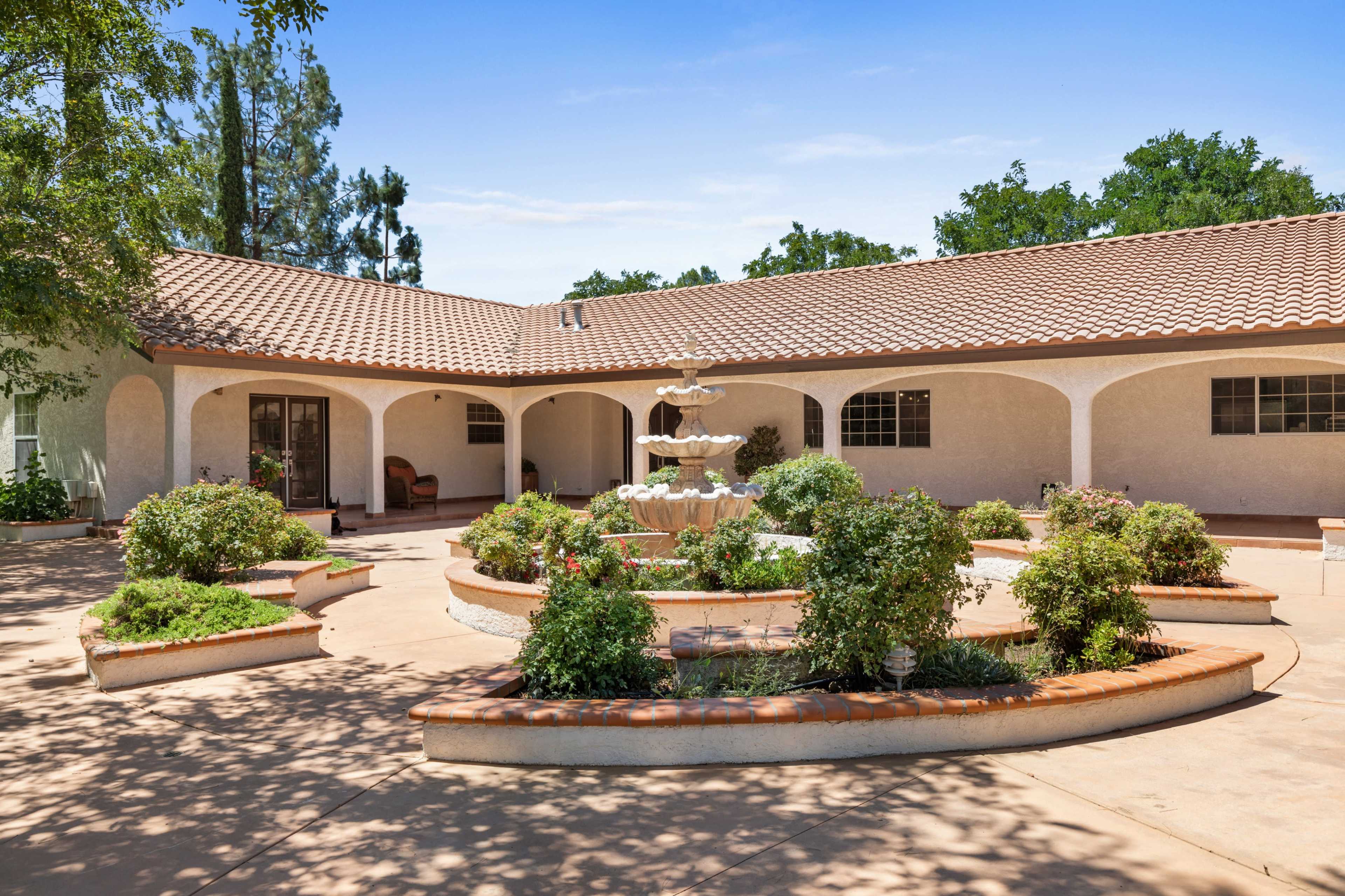 The image shows a courtyard with a three-tiered fountain surrounded by landscaped garden beds and a single-story building in the background.