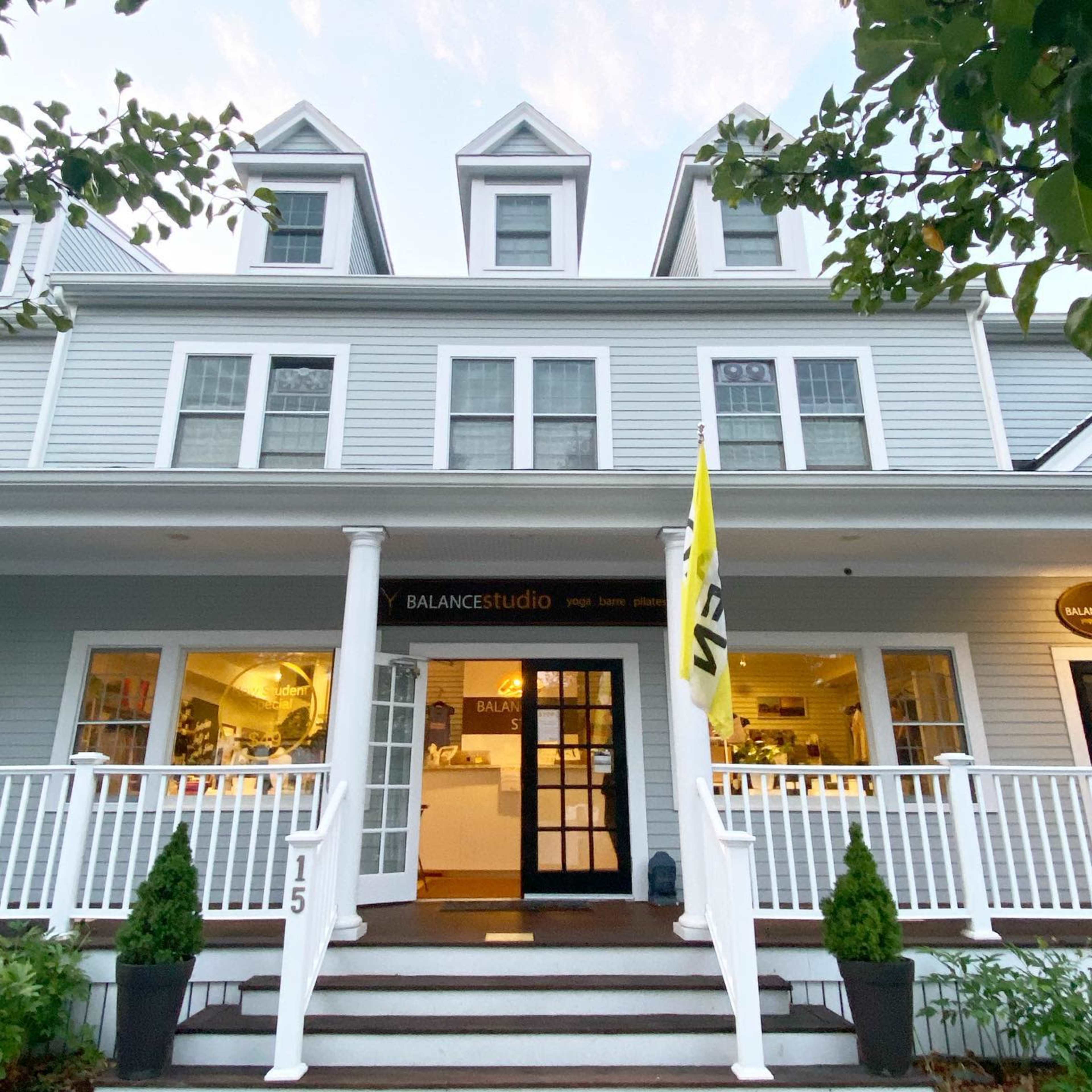 A two-story house with a porch, featuring a sign for "BALANCEstudio" and a yellow flag, surrounded by small shrubs.