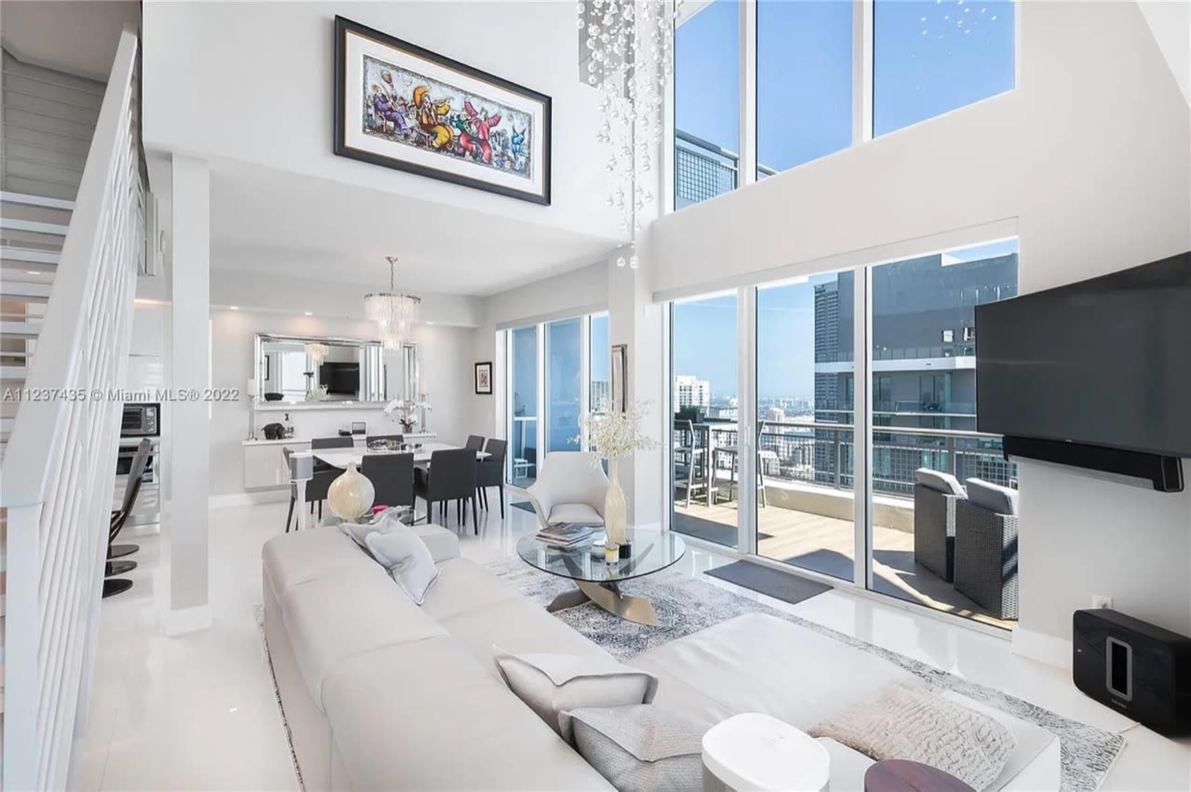 A modern living room and dining area with large windows, white furnishings, and a balcony overlooking a city skyline.