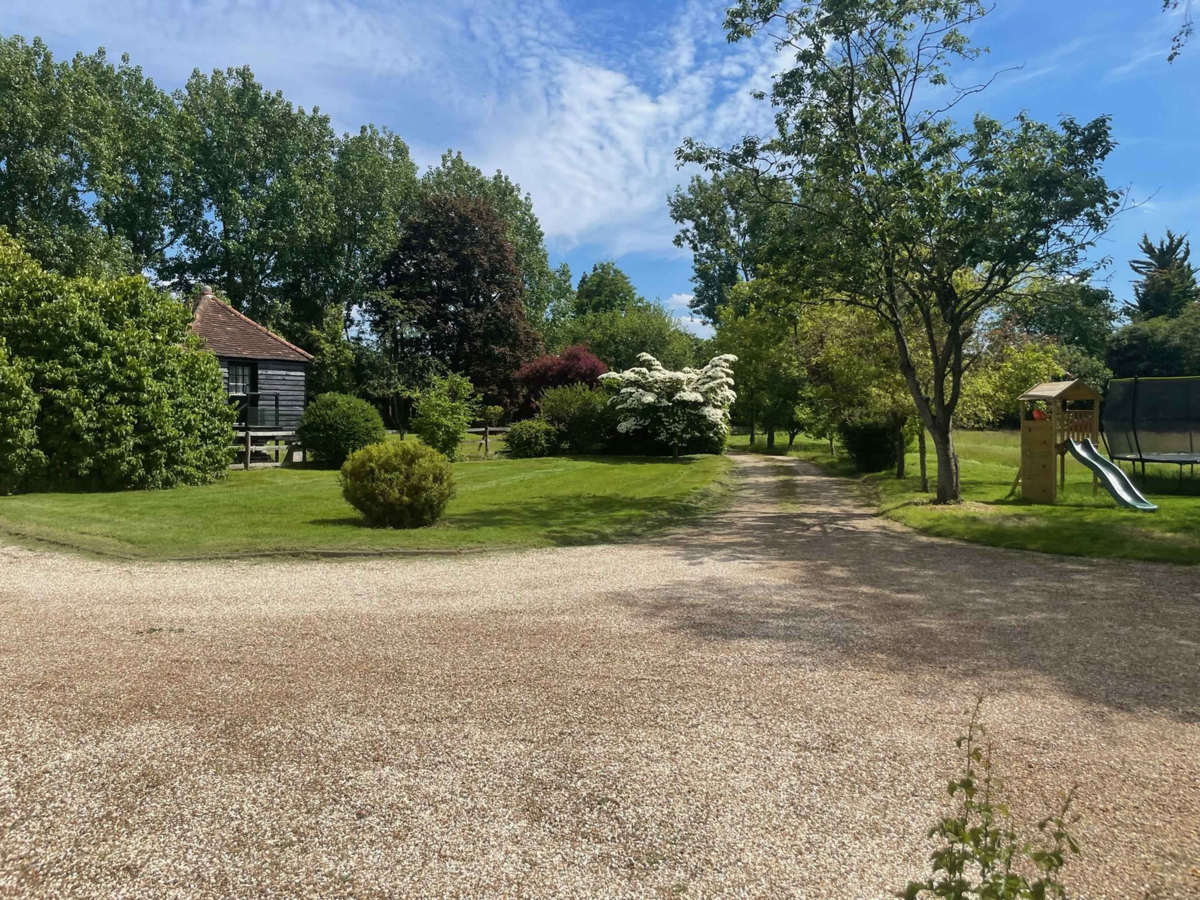 A gravel path leads through a green landscape with a wooden building, trees, and a children's slide in the background.