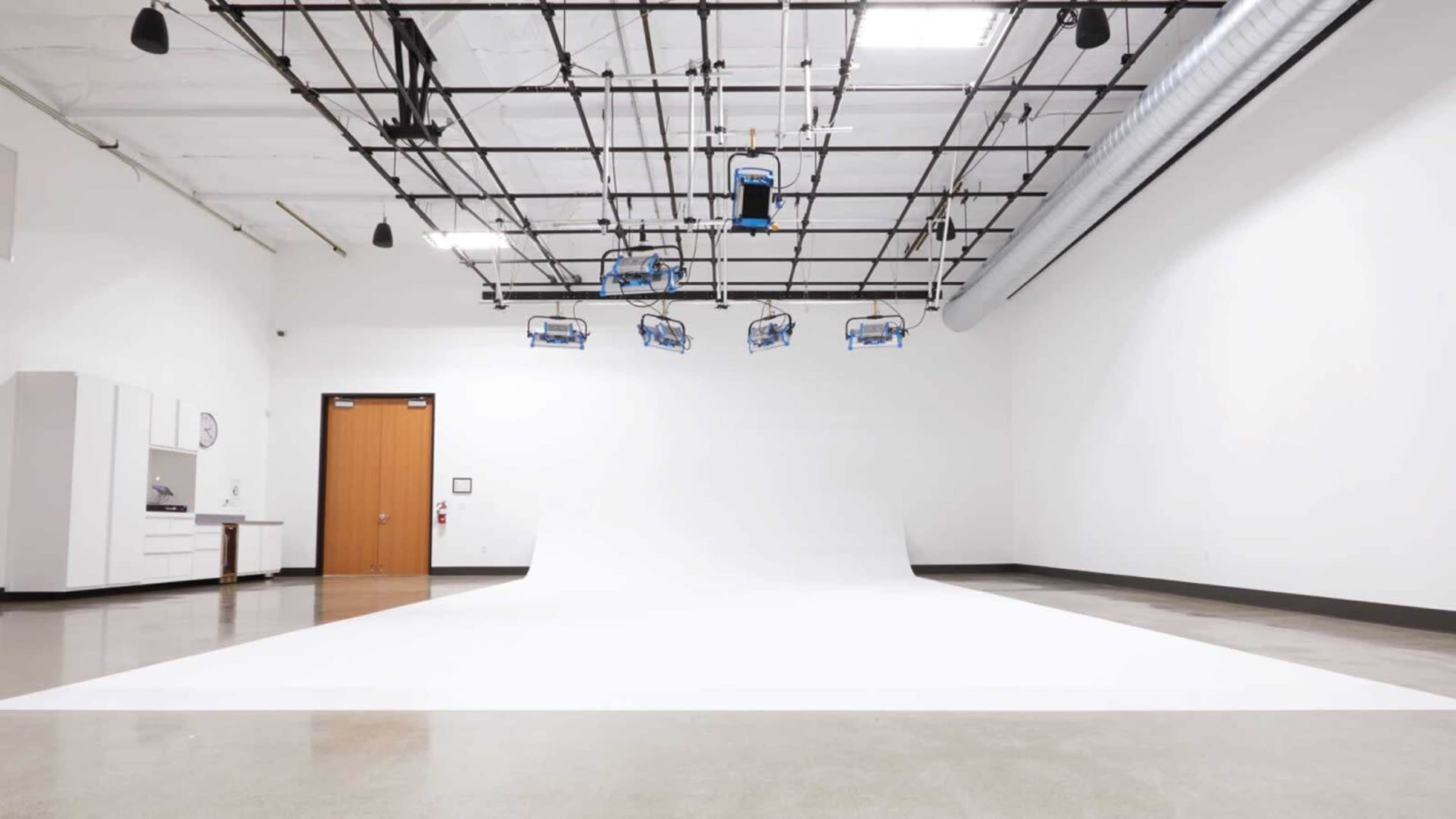 The image shows a spacious, empty studio with a white backdrop, overhead lighting fixtures, and a closed door in the background.