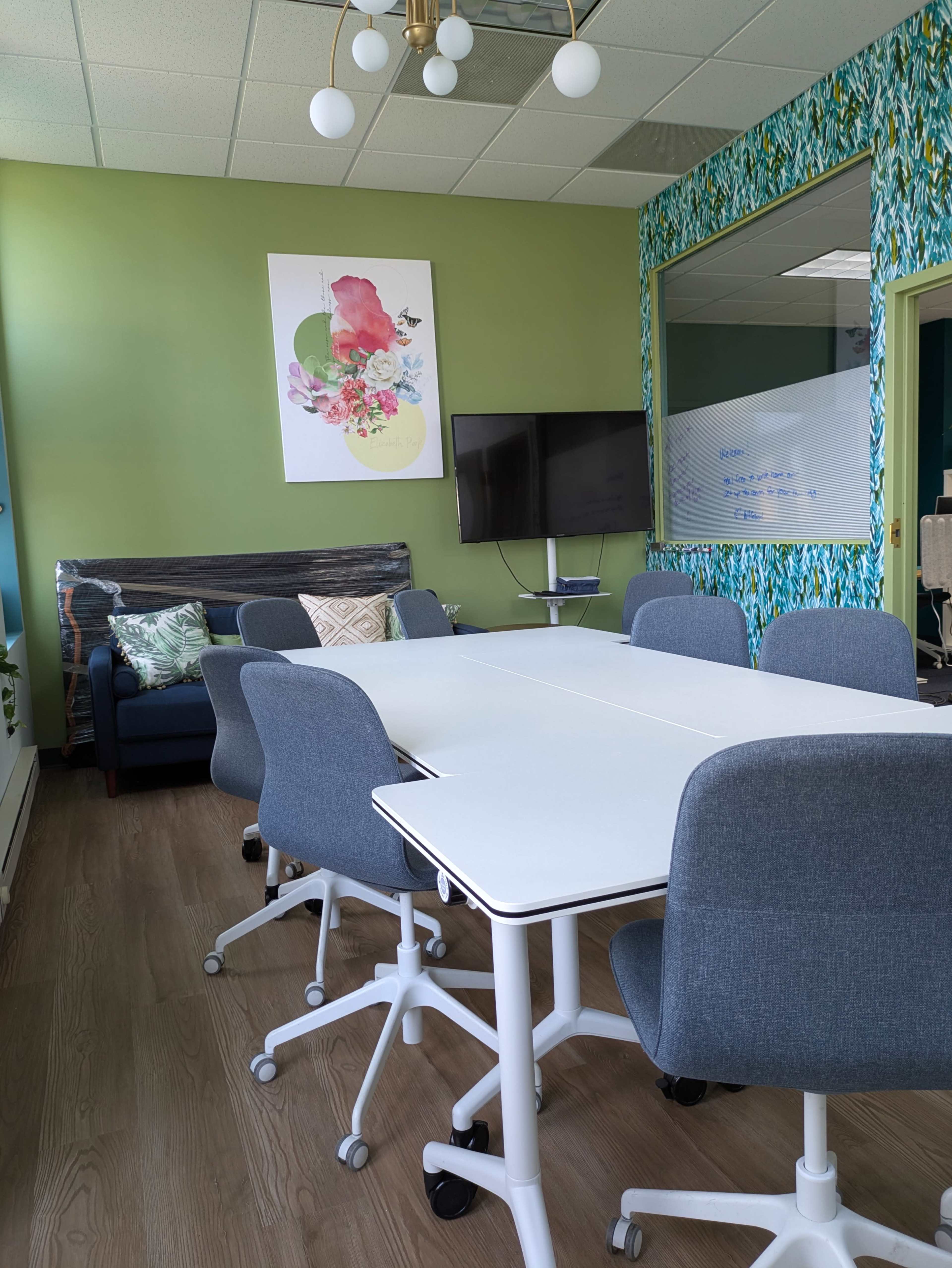 The image shows a modern conference room with a large table surrounded by grey office chairs, a sofa in the corner, a television mounted on the wall, and colorful wallpaper.