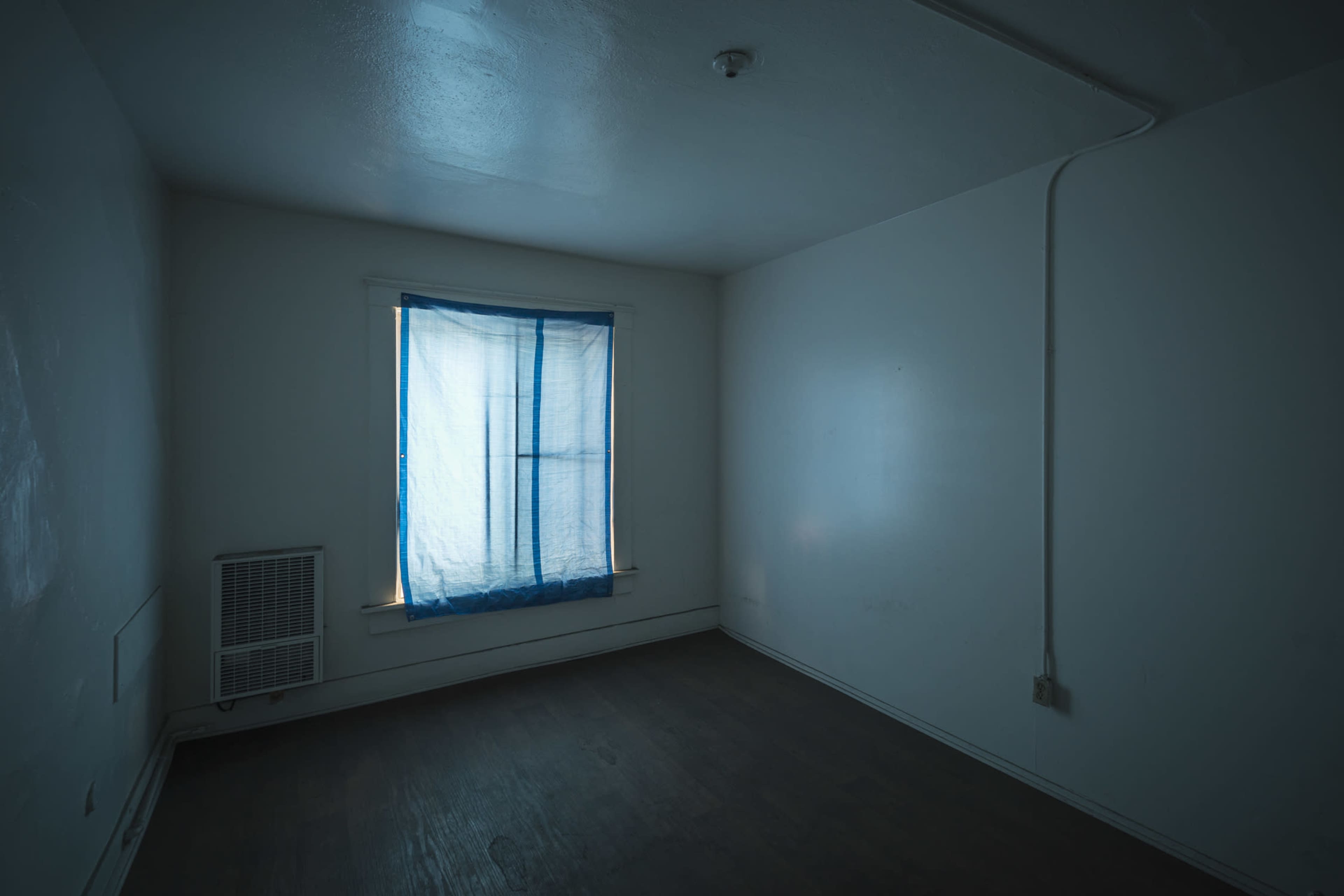 A sparse room with bare walls, a wooden floor, and a window covered by a blue curtain.