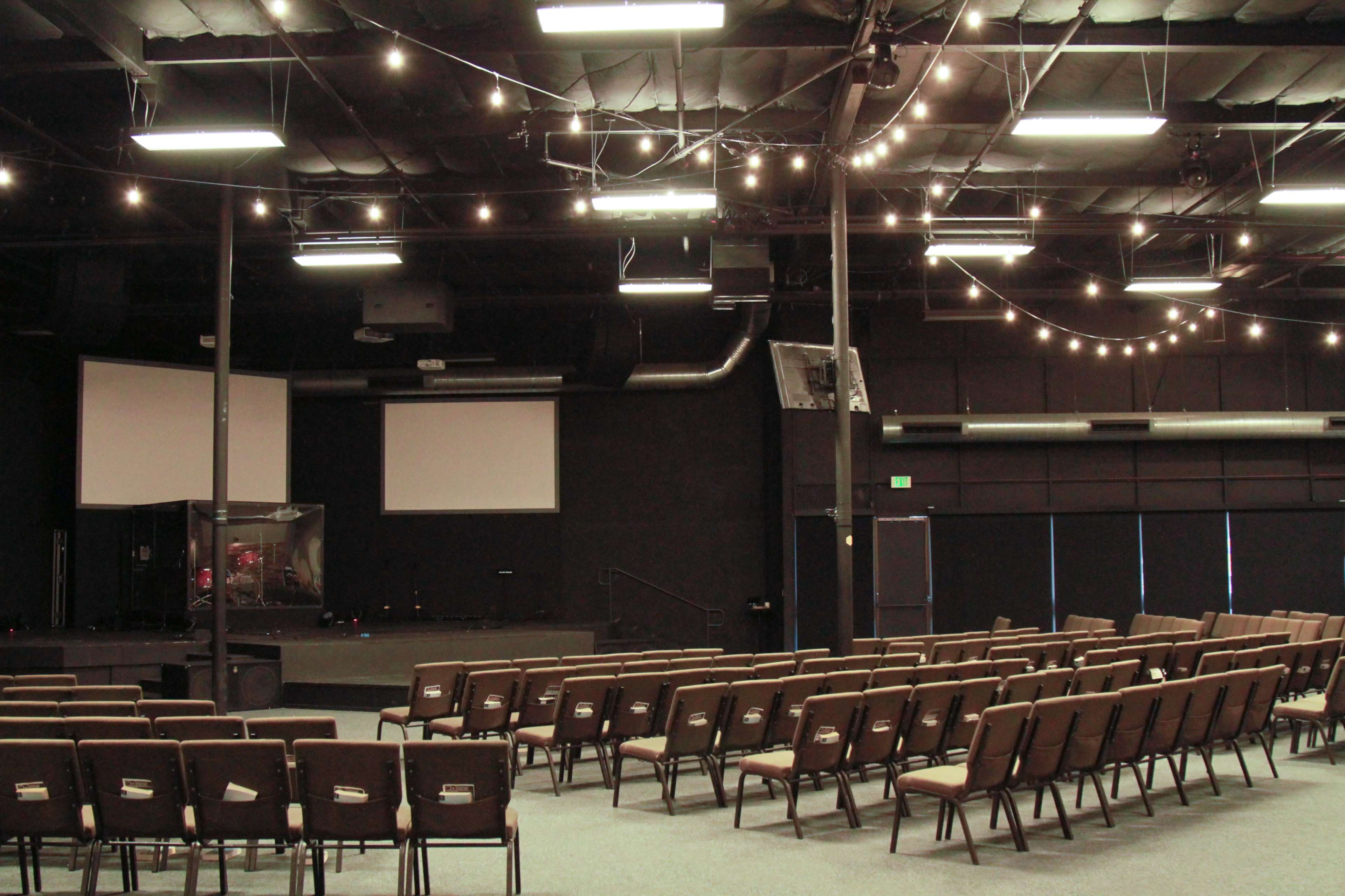 The image shows a dimly lit auditorium with rows of chairs facing a stage that has two blank projection screens.