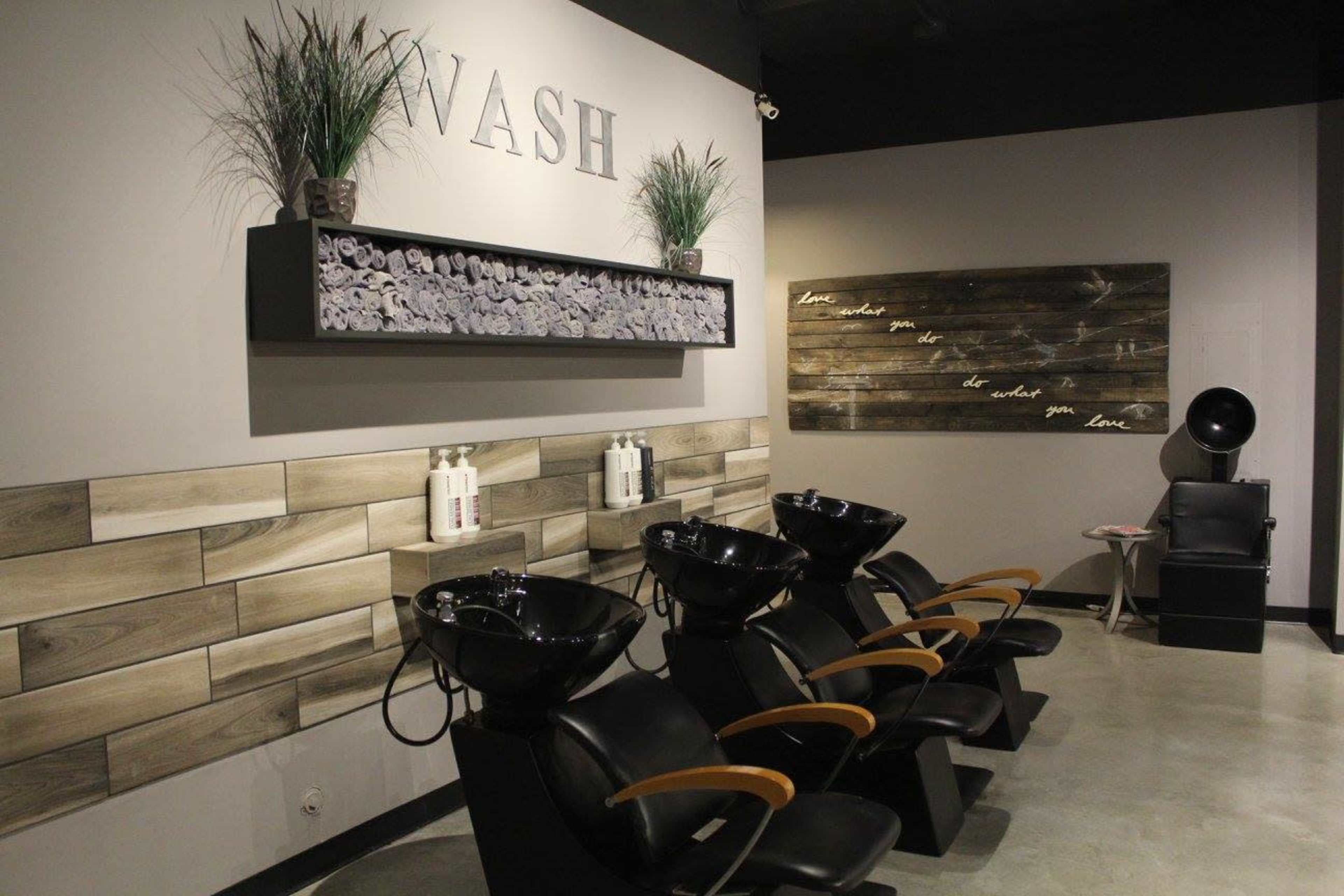 The image shows a hair salon interior featuring several black shampoo chairs arranged in front of a wall with a decorative shelf displaying rolled towels and hair care products.
