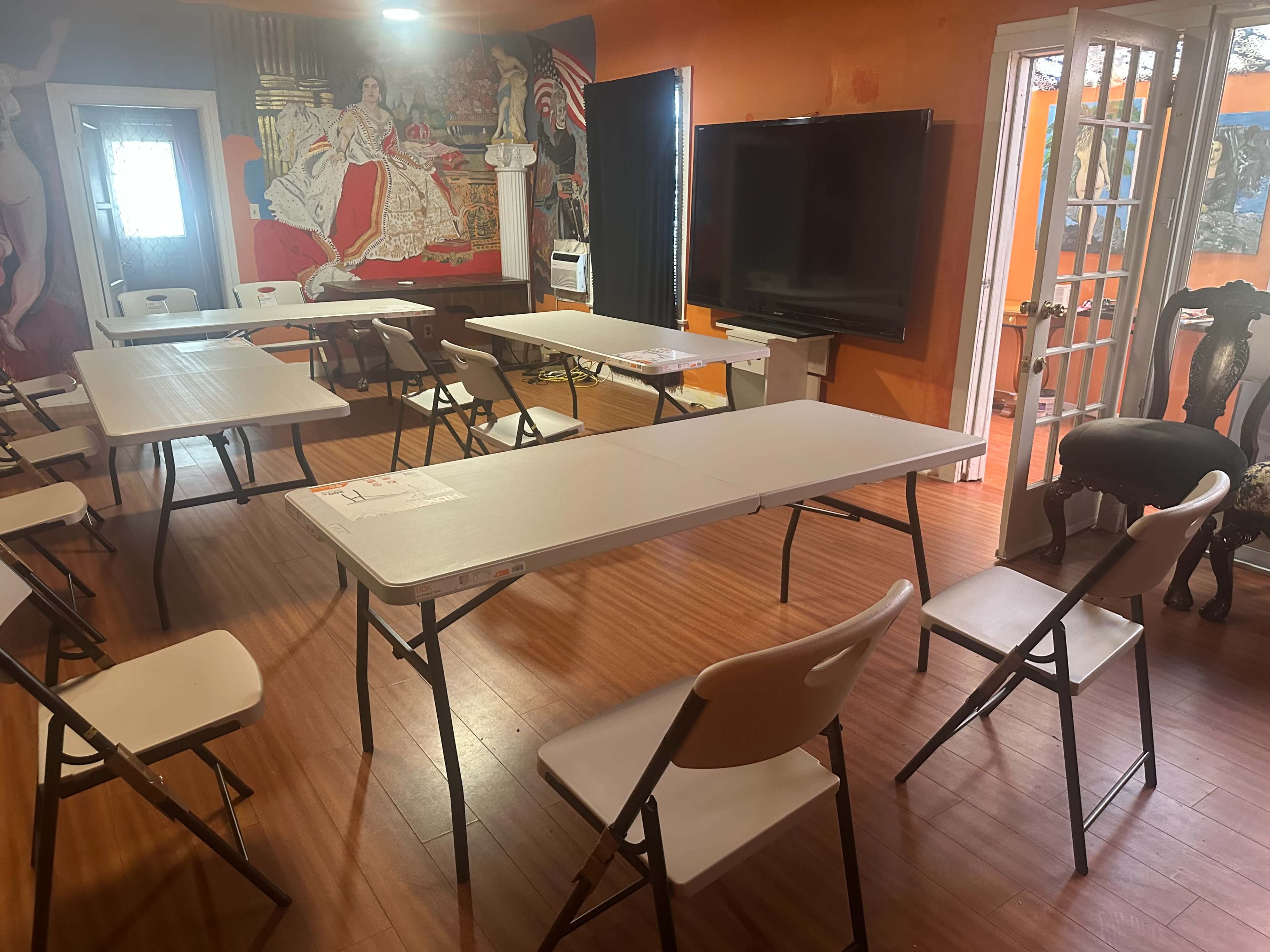 A room with orange walls contains several folding tables and chairs arranged for a meeting, along with a large television and a mural on one wall.