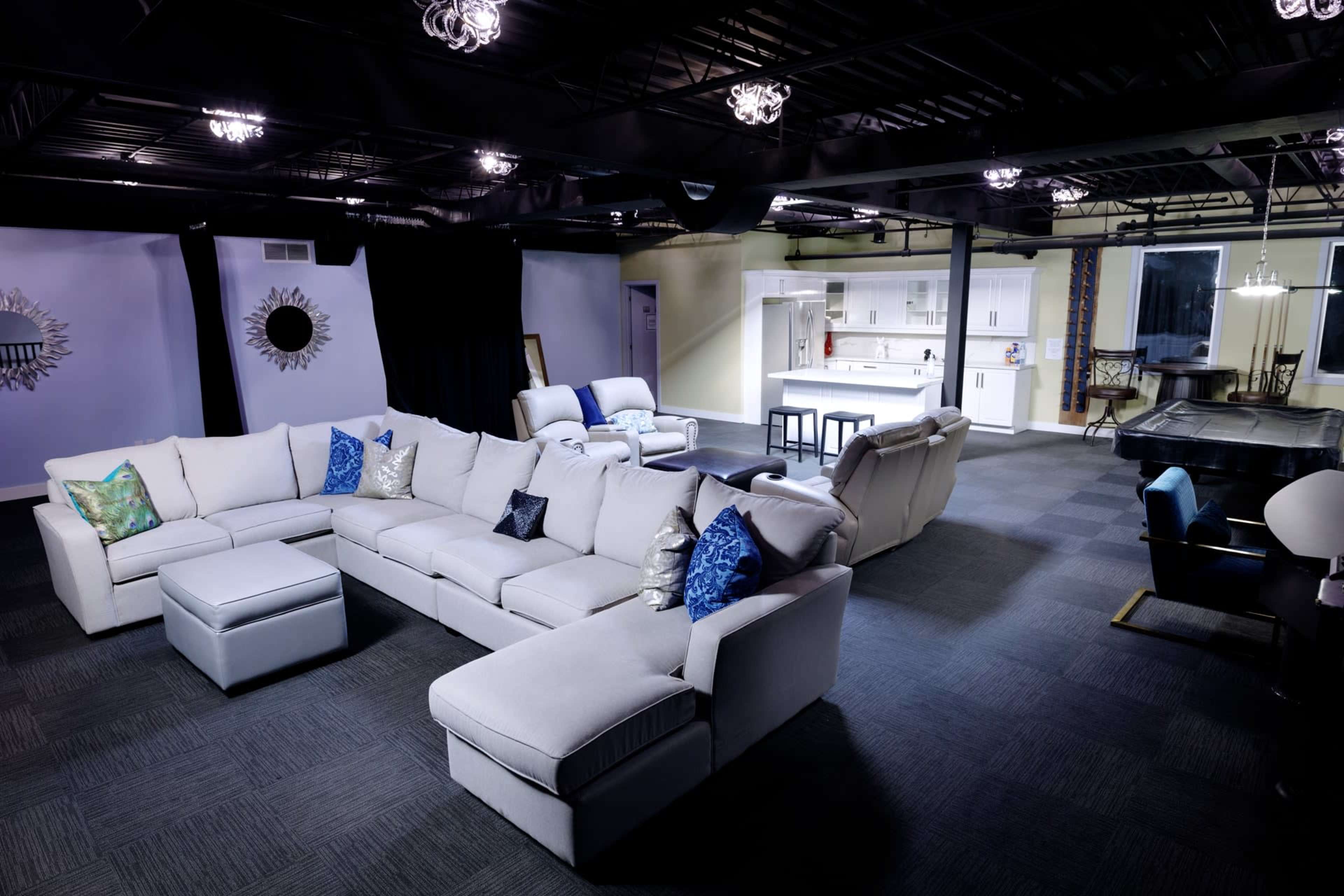 A spacious living area featuring a large sectional sofa with decorative pillows, a minimalist dining set, and a separate kitchen area with white cabinets in the background.