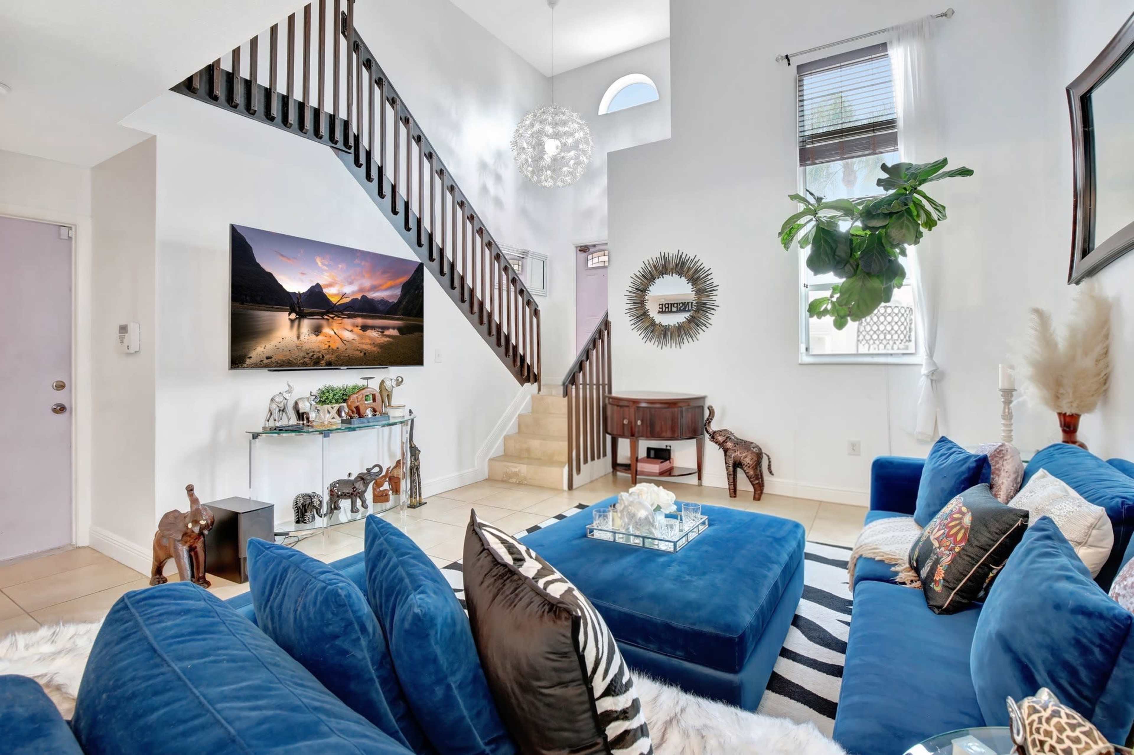 A stylish living room features a large blue sectional sofa, a glass coffee table, and a staircase leading to an upper level, with decorative plants and a large TV mounted on the wall.